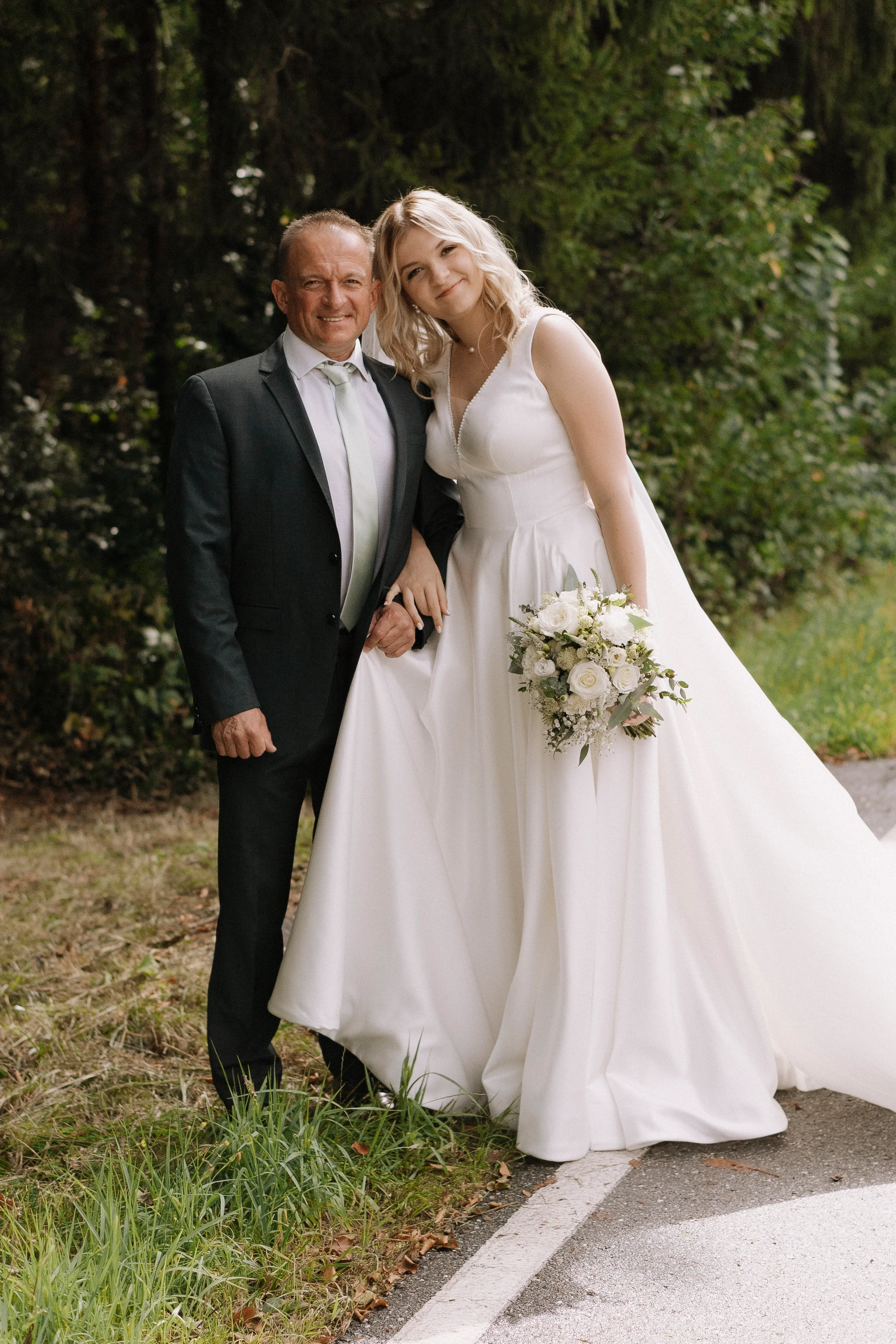 Ein älterer Mann und eine junge Frau in Hochzeitskleidung stehen nebeneinander im Freien, umgeben von Bäumen, und lachen. Die Frau hält einen Brautstrauß, der aus weißen Rosen und grünem Laub besteht.