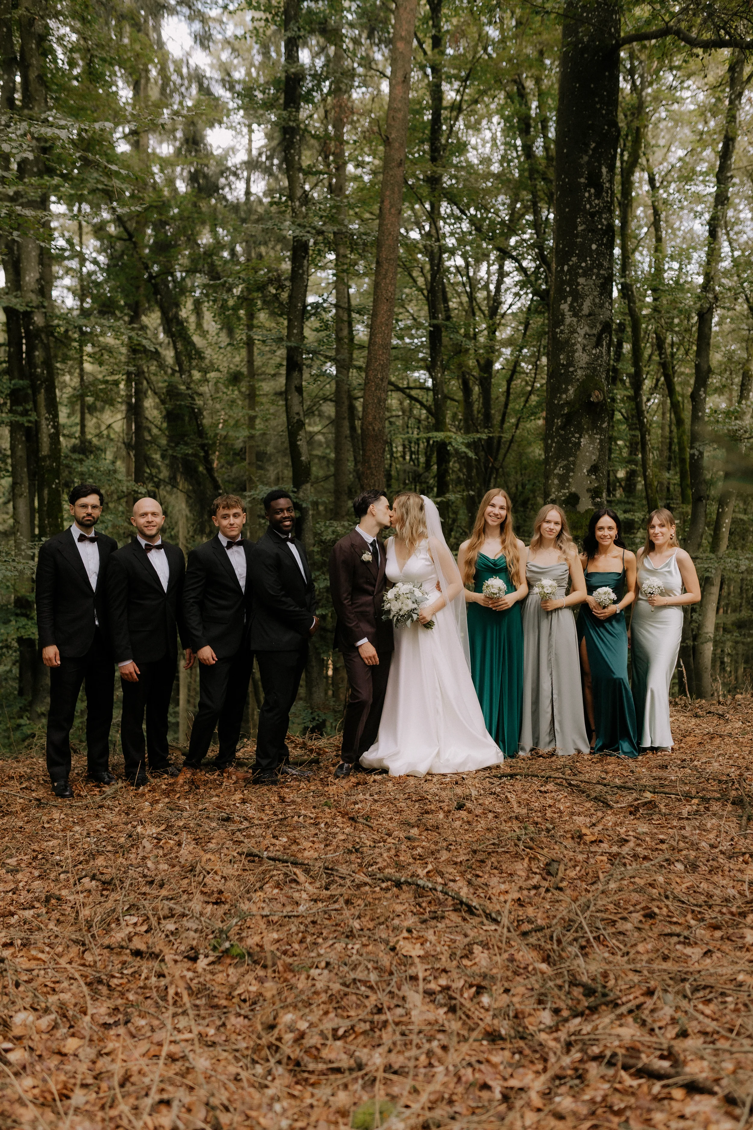 Eine Gruppe von Hochzeitsgästen steht in einem Wald, vier Frauen in eleganten Abendkleidern und vier Männer in Anzügen, die Braut und der Bräutigam, die sich küssen, in der Mitte. Die Naturkulisse besteht aus hohen Bäumen und Herbstlaub auf dem Boden