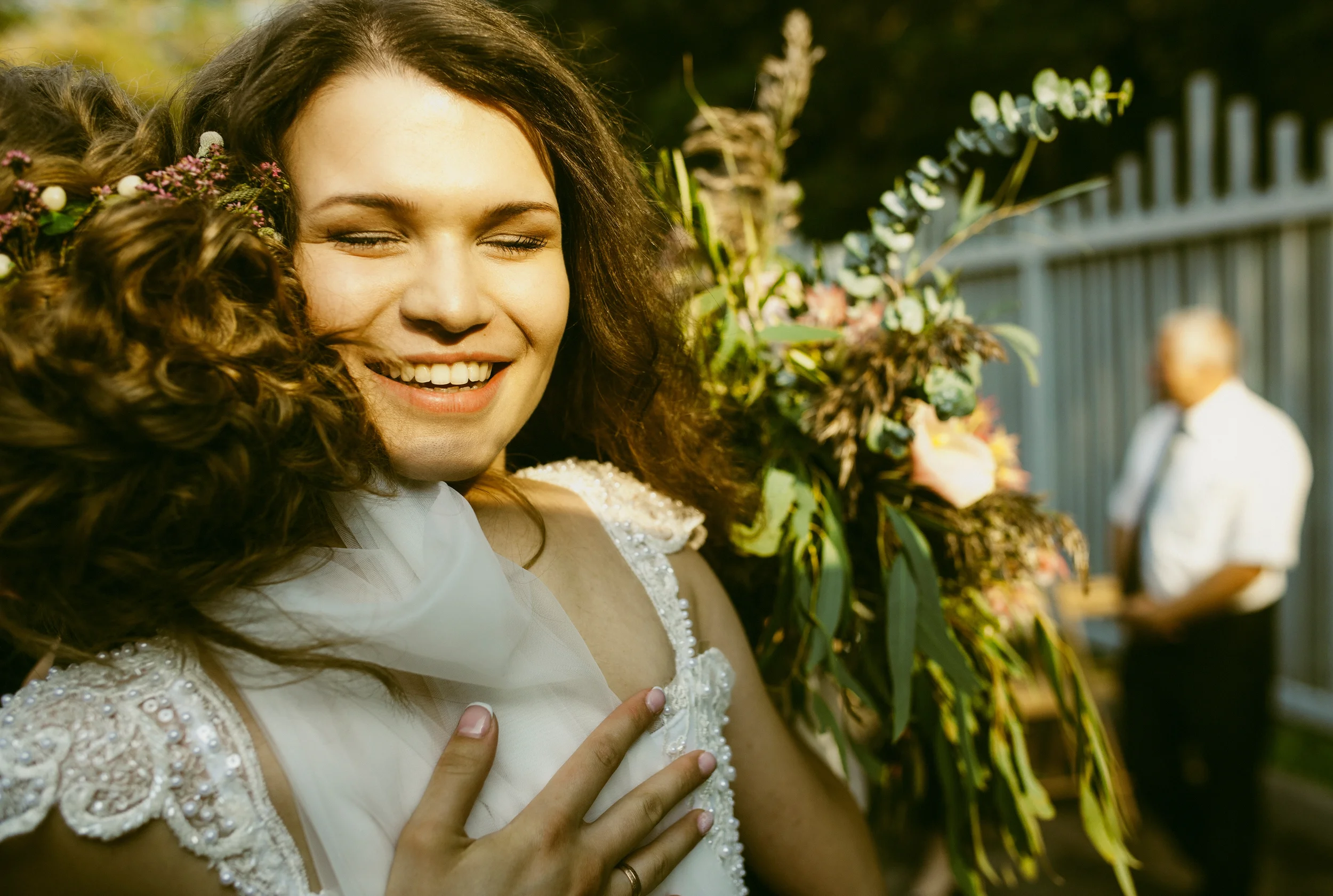 Junge Frau bei einer Hochzeit hält ihre Hand auf ihre Brust, um die Hand ihrer Braut zu berühren, im Hintergrund sind Blumen und ein Zaun sichtbar.