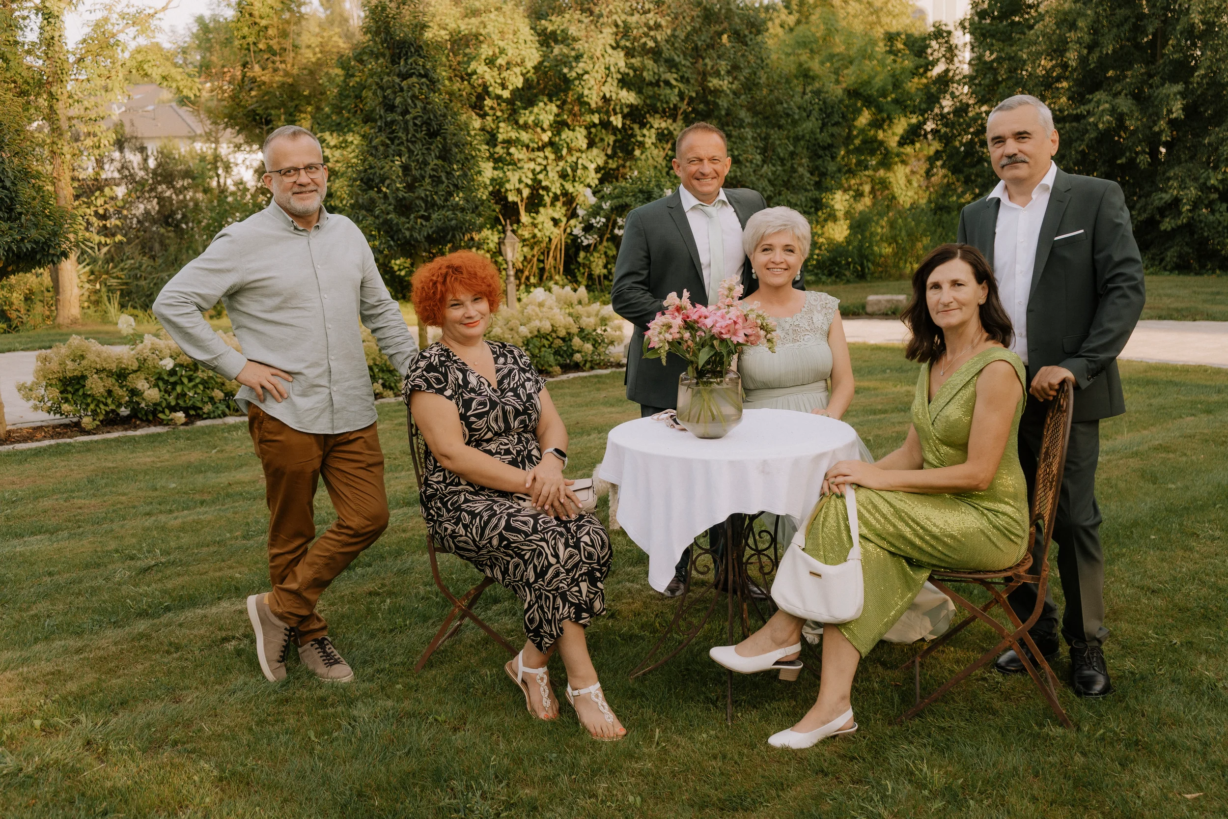 Eine Gruppe von sieben Menschen posiert im Garten bei Sonnenuntergang, einige sitzen um einen kleinen runden Tisch mit Blumen, andere stehen dahinter. Alle tragen elegante Kleidung.