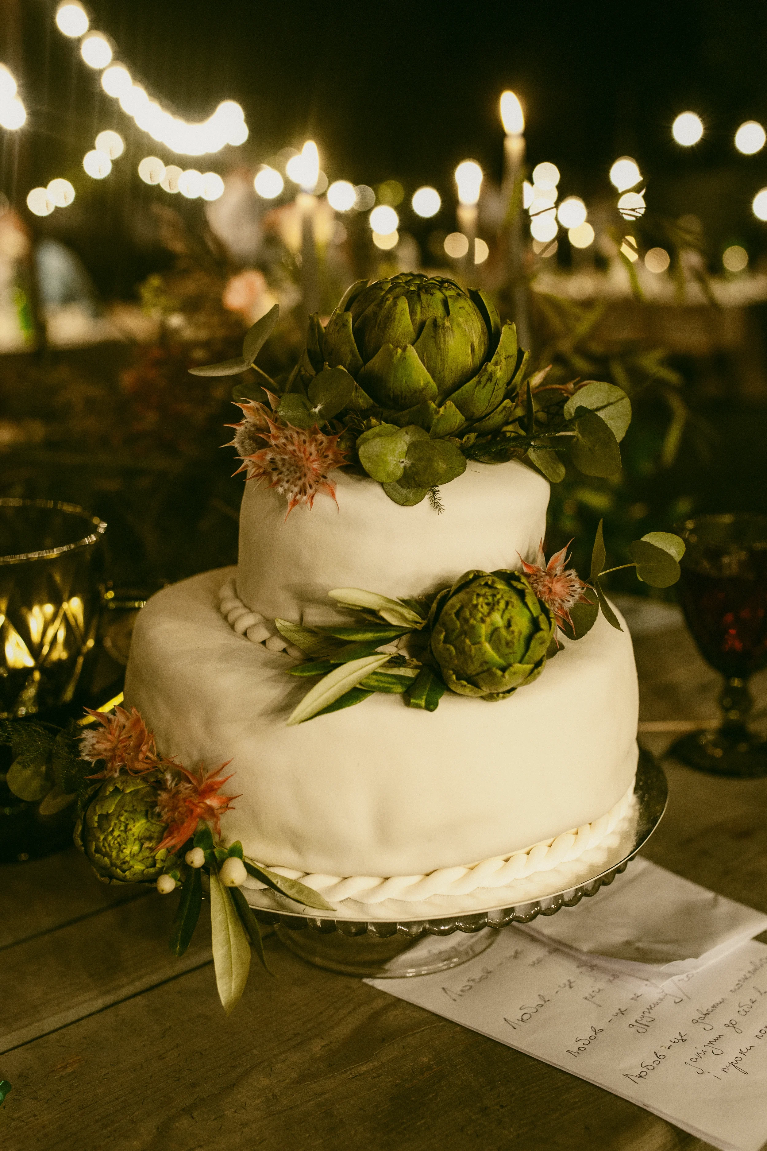 Ein zweistöckiger, eleganter weißer Hochzeitstorte mit grünen Artischocken und Blumen als Dekoration, auf einem silbernen Tortenständer, im Hintergrund Lichter bei Nacht.
