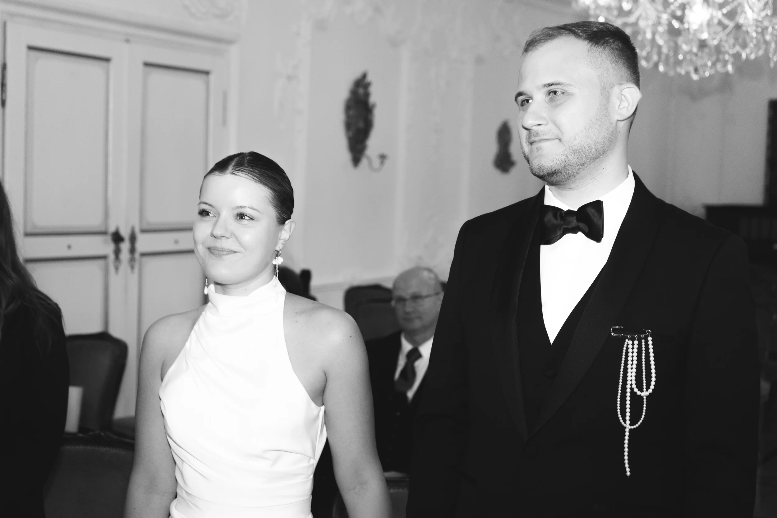 Schwarz-weiß Foto eines Mannes und einer Frau bei einer formellen Veranstaltung, beide in eleganten Kleidern. Der Mann trägt einen schwarzen Anzug mit Fliege und Perlenkette, die Frau trägt ein weißes Kleid mit hohem Kragen und langen Ohrringen.