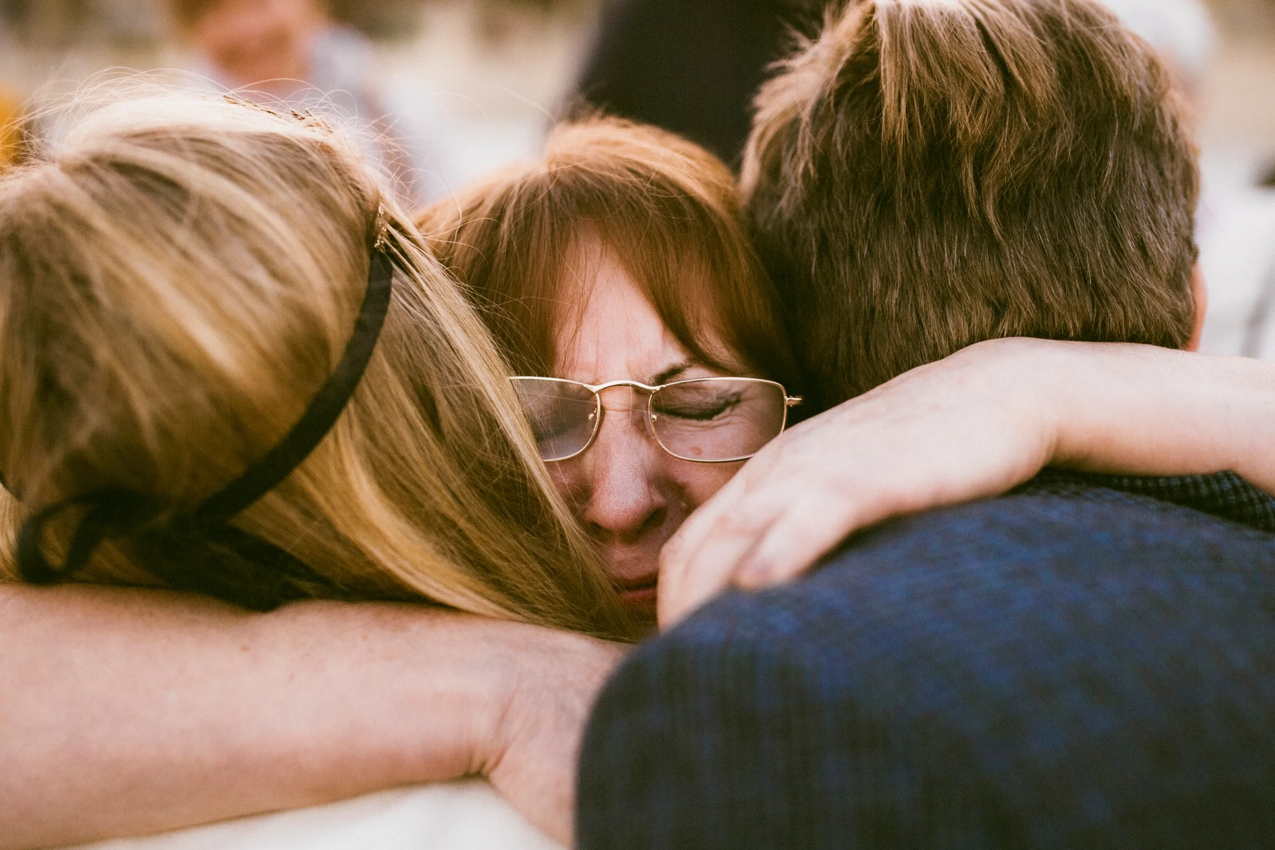 Mehrere Menschen umarmen sich herzlich, eine Person trägt Brille und hat rote Haare, es ist eine emotionale Umarmung.