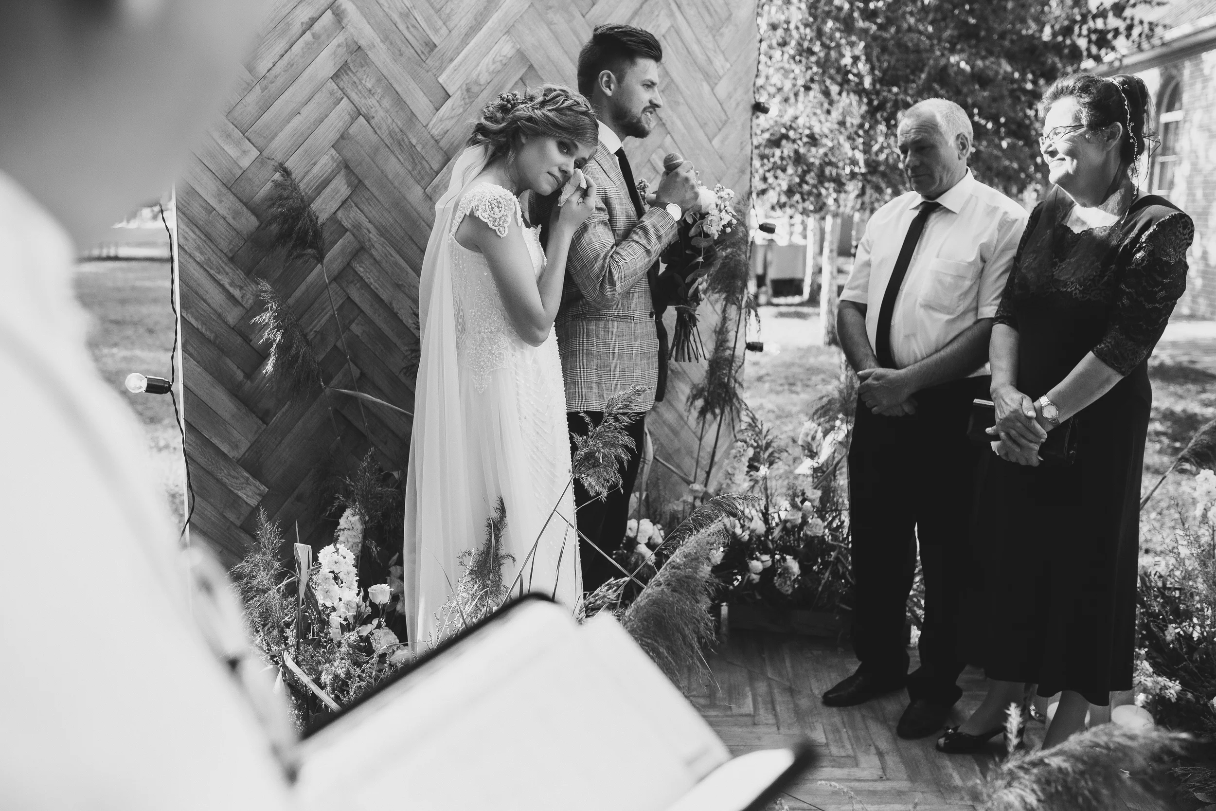 Schwarz-weiß-Foto einer Hochzeit mit Braut in Weiß und Bräutigam in Anzug, die vermutlich während einer Zeremonie sprechen, umgeben von drei Erwachsenen, bei einem Fest im Freien.