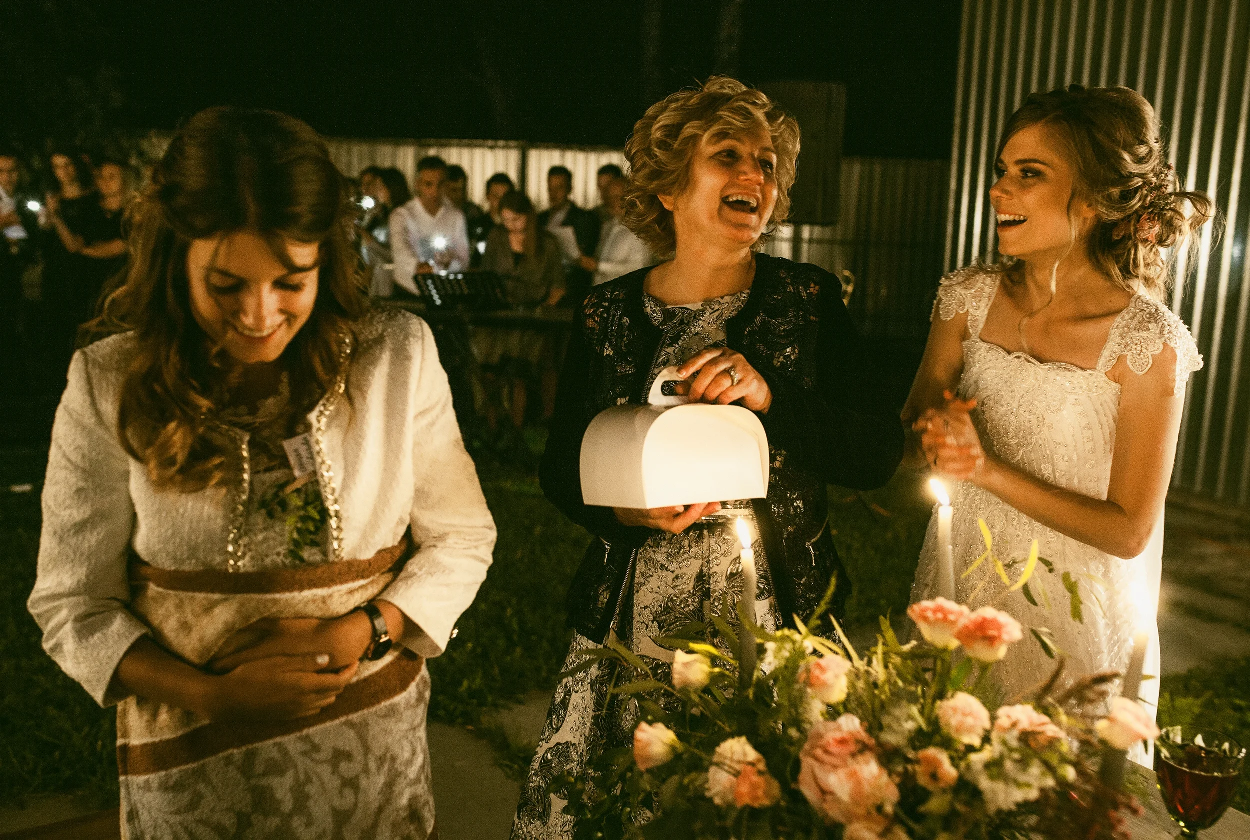 Mehrere Frauen feiern bei einem Abendessen im Freien mit Kerzen und Blumen, lachen und unterhalten sich festlich gekleidet bei Nacht.