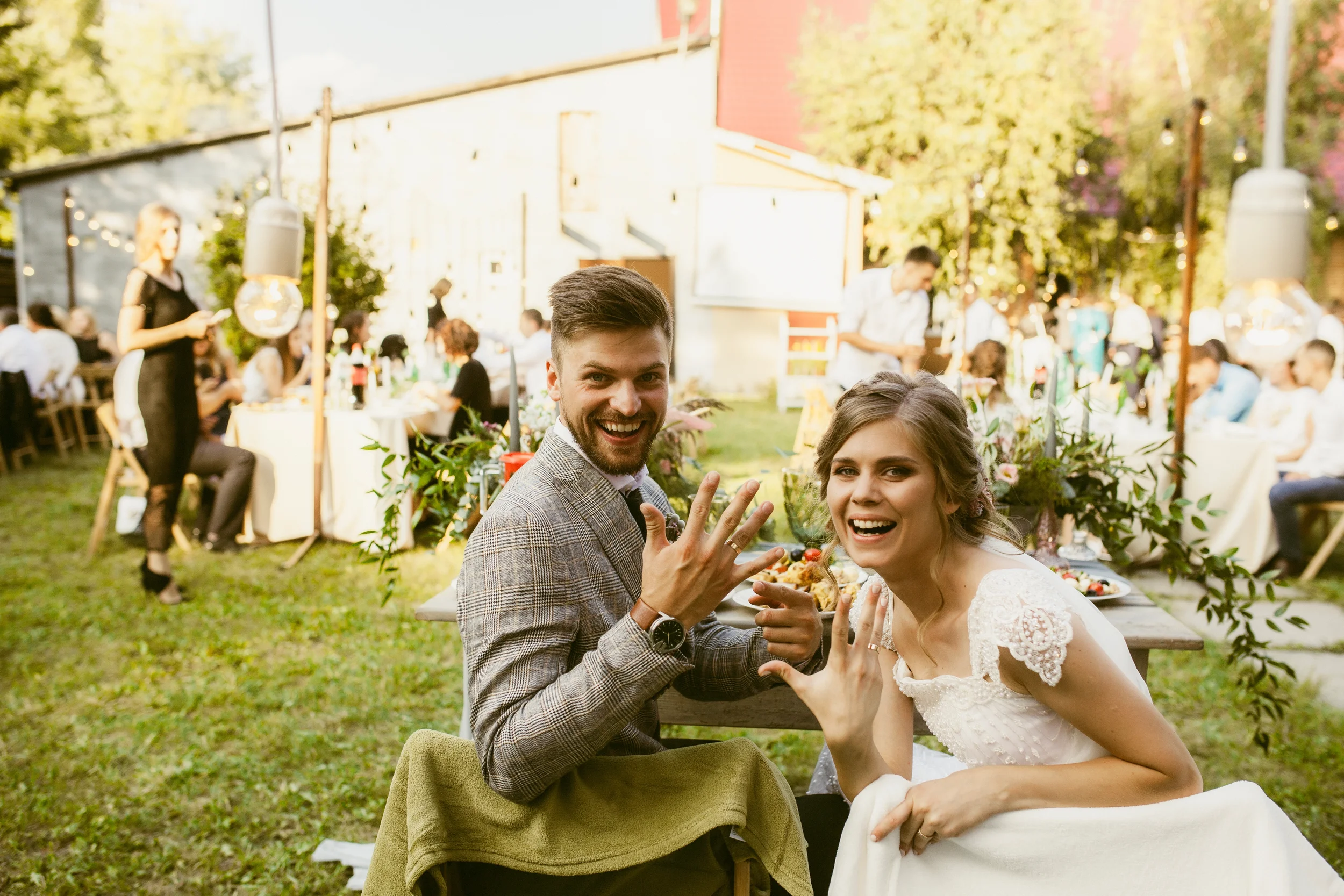 Ein lachendes Paar, wahrscheinlich Braut und Bräutigam, bei einer Gartenparty im Freien, umgeben von anderen Gästen, die an einem festlich gedeckten Tisch sitzen, mit viel Grün und warmem Sonnenlicht im Hintergrund.