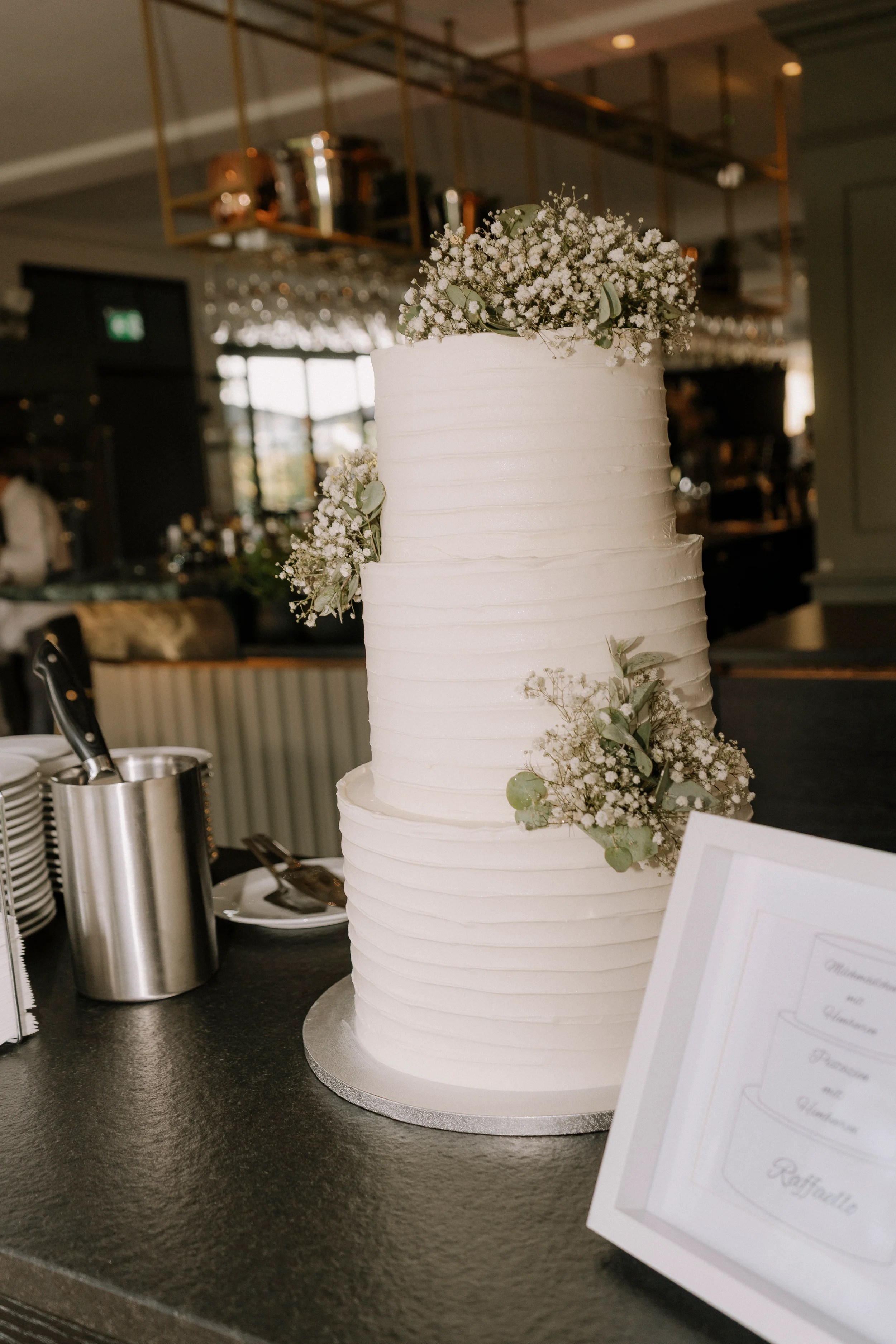 Ein mehrstöckiger weißer Hochzeitstorte mit Blumen, auf einem schwarzen Tisch in einem Restaurant oder Veranstaltungsraum.