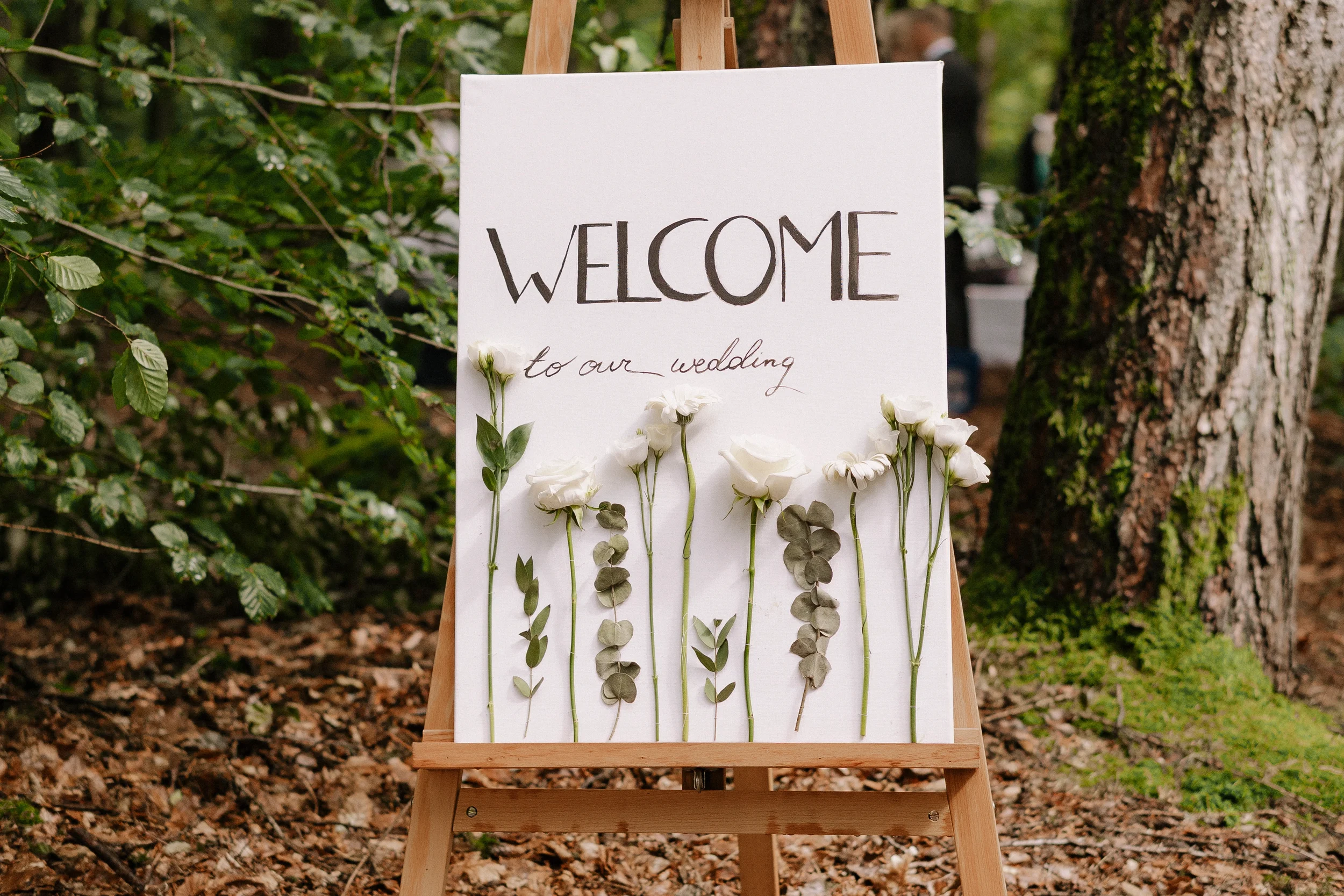 Ein Willkommensschild für eine Hochzeit, das auf einem Holzständer im Freien steht. Das Schild ist weiß mit handgeschriebenem Text "WELCOME to our wedding" und ist mit weißen Blumen dekoriert.