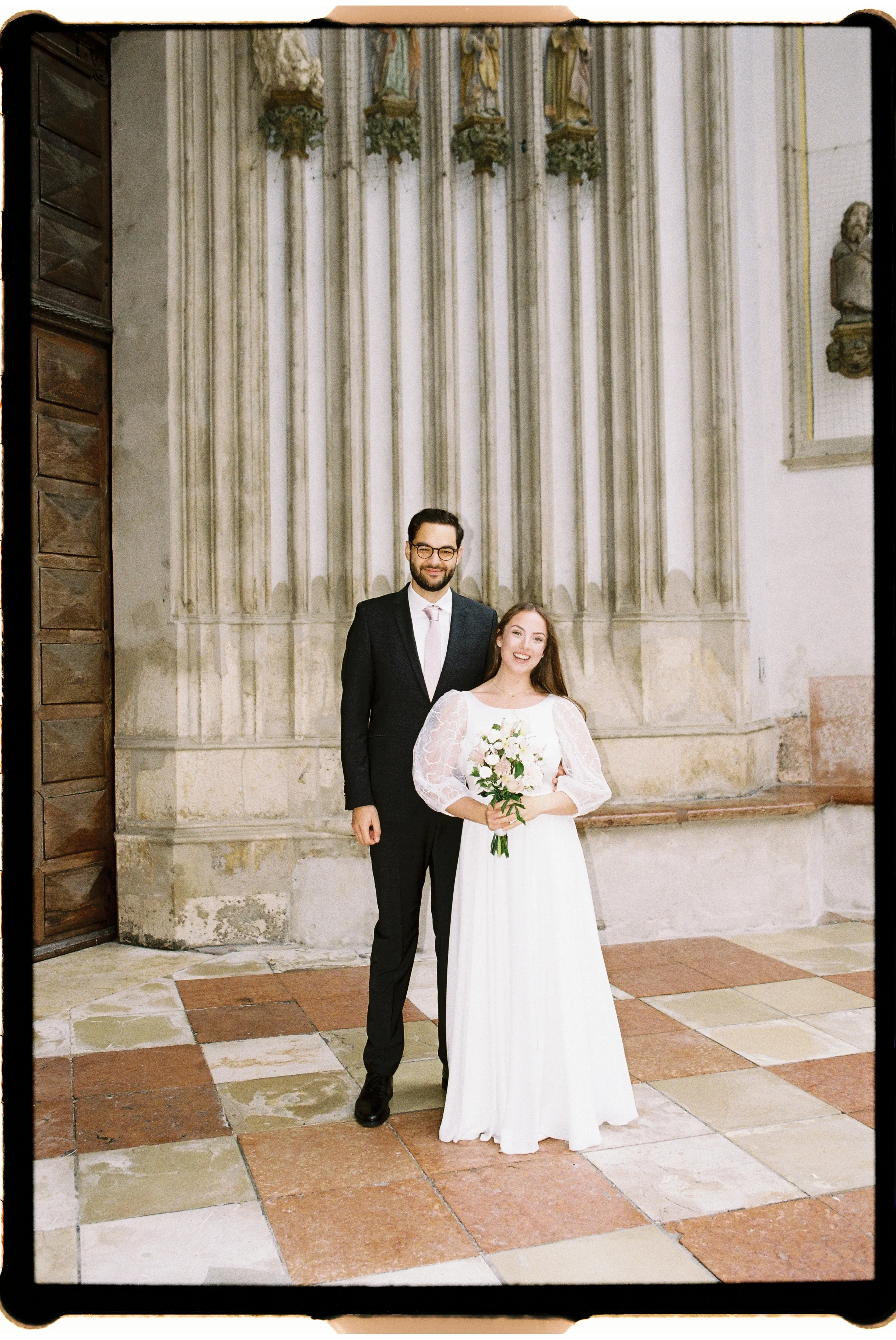 Ein verheiratetes Paar während ihrer Hochzeitszeremonie in einer Kirche, die Frau trägt ein weißes Brautkleid und hält einen Blumenstrauß, der Mann trägt einen schwarzen Anzug mit Krawatte.