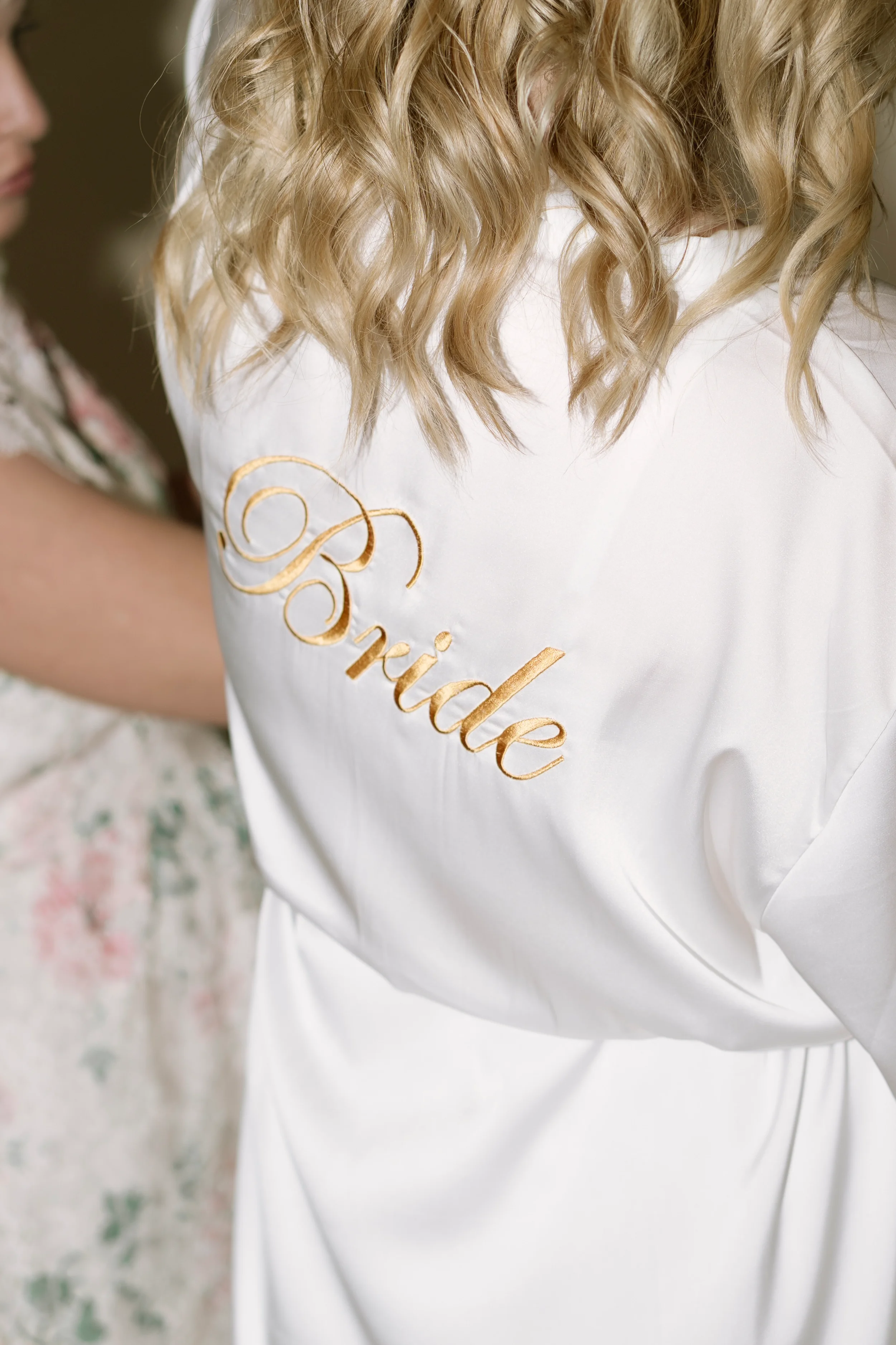 Nahaufnahme eines weißen Hochzeitskleides mit goldener Stickerei auf der Schulter und lockigem, blonden Haar. Im Hintergrund ein Blumentapete.