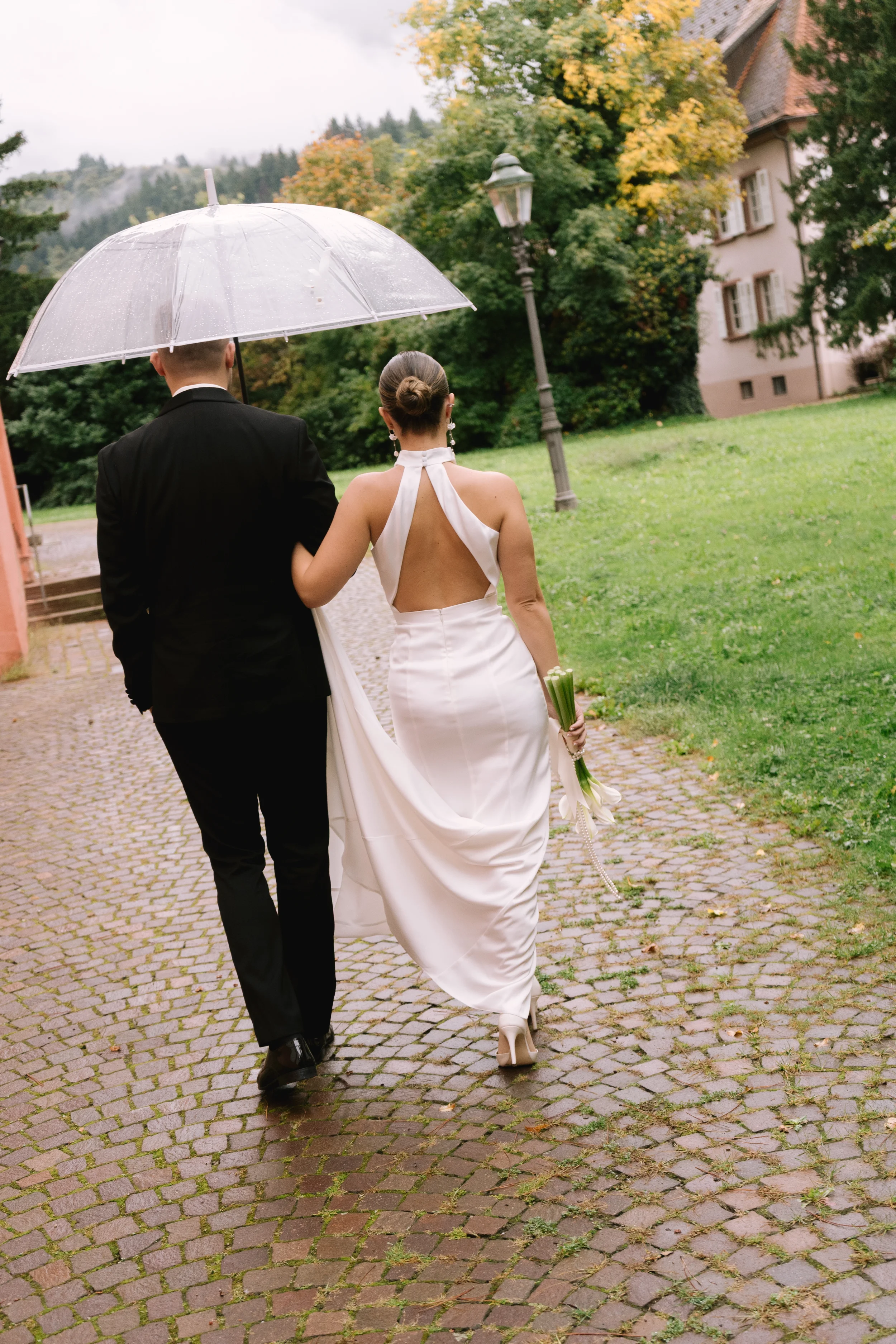Ein Paar bei einer Hochzeitszeremonie, das sich anmutig auf einem gepflasterten Weg in einer grünen Parklandschaft mit Bäumen und einem alten Haus im Hintergrund bewegt, während es einen transparenten Regenschirm hält.