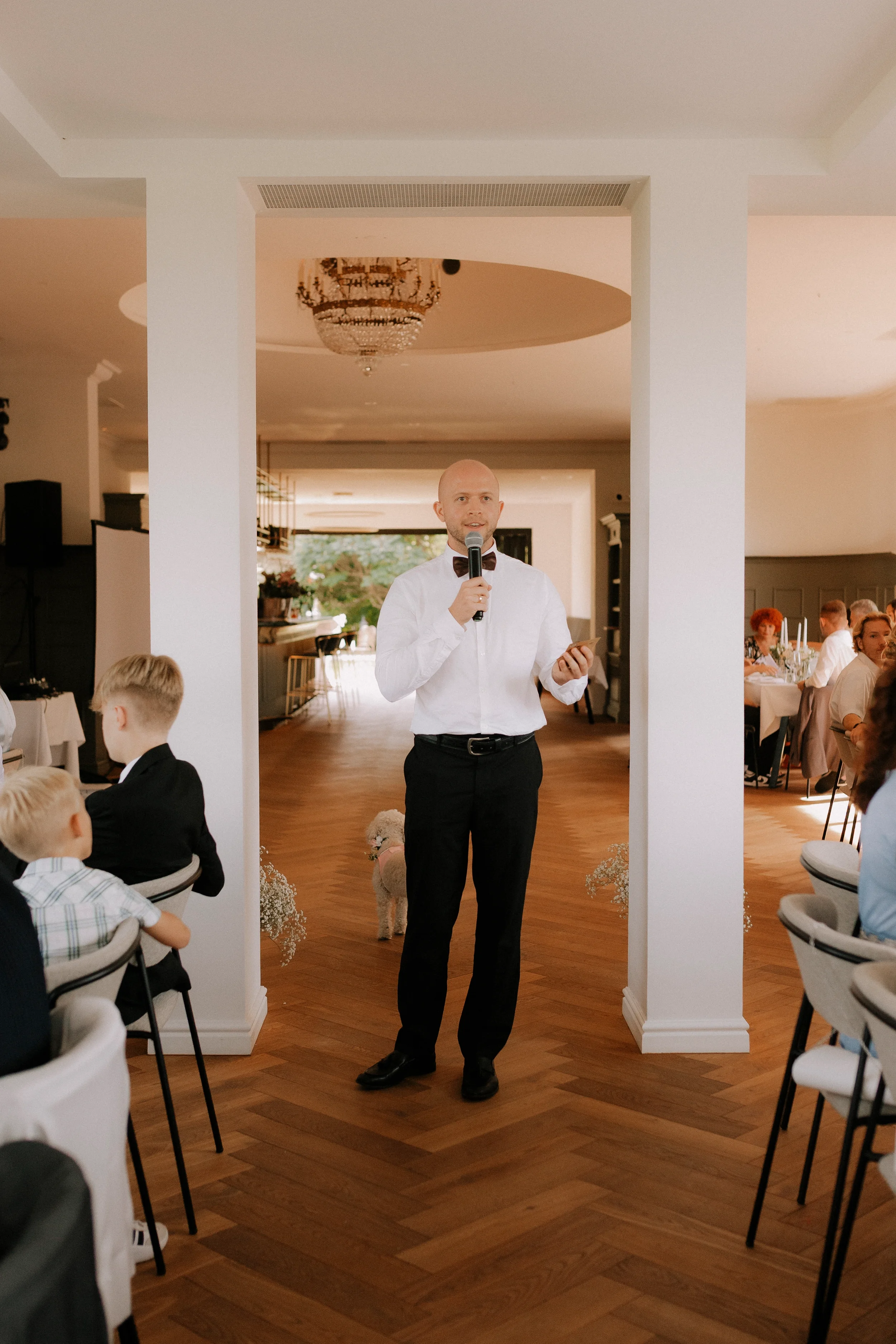 Mann in Weißem Hemd und Fliege hält Mikrofon und spricht bei einer Feier. Im Hintergrund sitzen Gäste an Tischen, es gibt eine elegante Decke mit Kronleuchter im Raum.