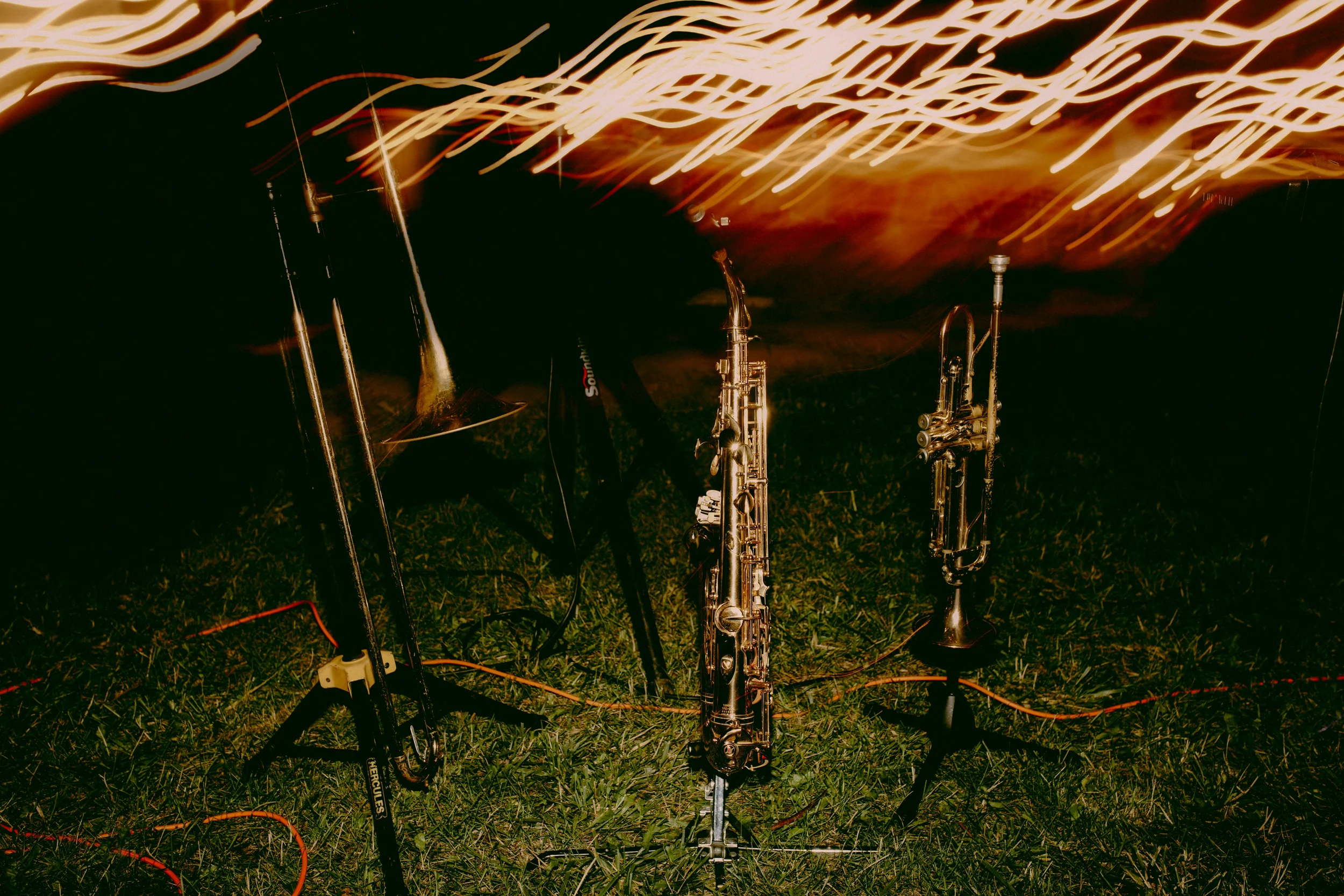 Mehrere Musikinstrumente, darunter Saxophone, sind nachts im Freien auf Gras aufgestellt, während bunte Lichtstreifen am Himmel leuchten.