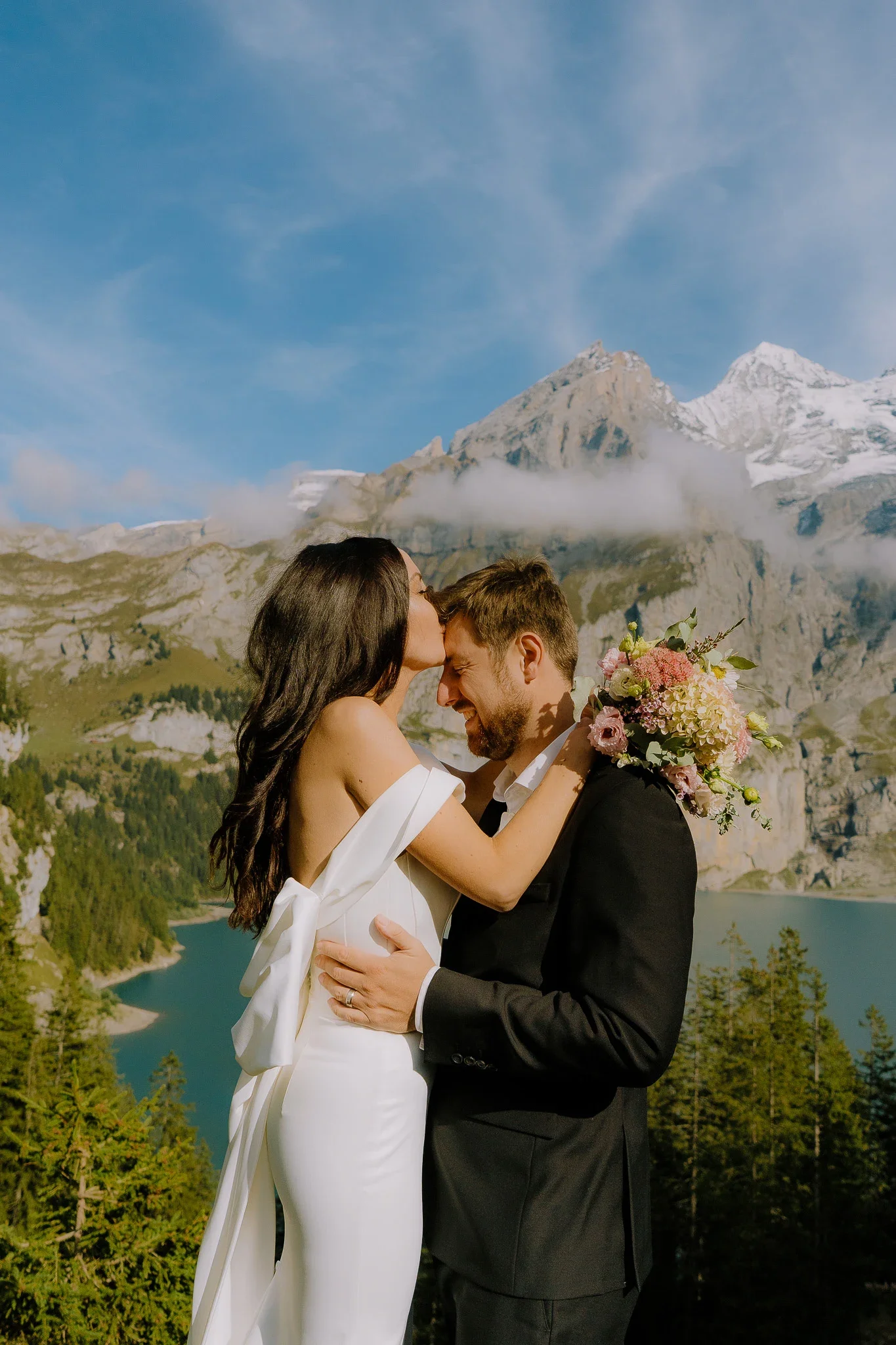 Ein Paar in Hochzeitskleidung, das sich küsst, vor einer Berglandschaft mit Gipfeln, Wäldern und einem See im Hintergrund.
