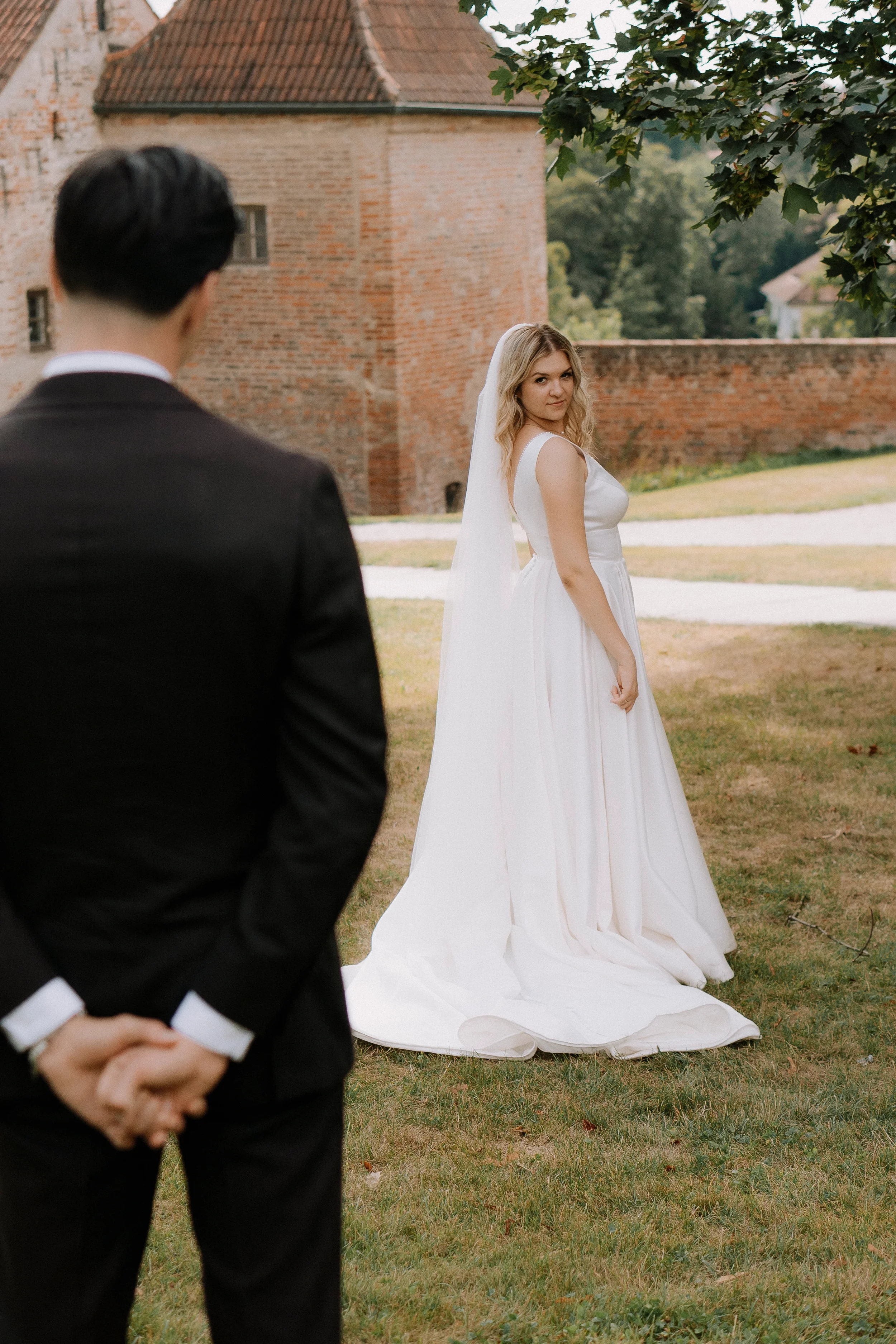 Ein Brautpaar bei einer Hochzeit im Freien vor einer alten Backsteinmauer, die Braut trägt ein weißes Hochzeitskleid mit Schleier, der Bräutigam trägt einen schwarzen Anzug, nur der Rücken des Bräutigams ist sichtbar.