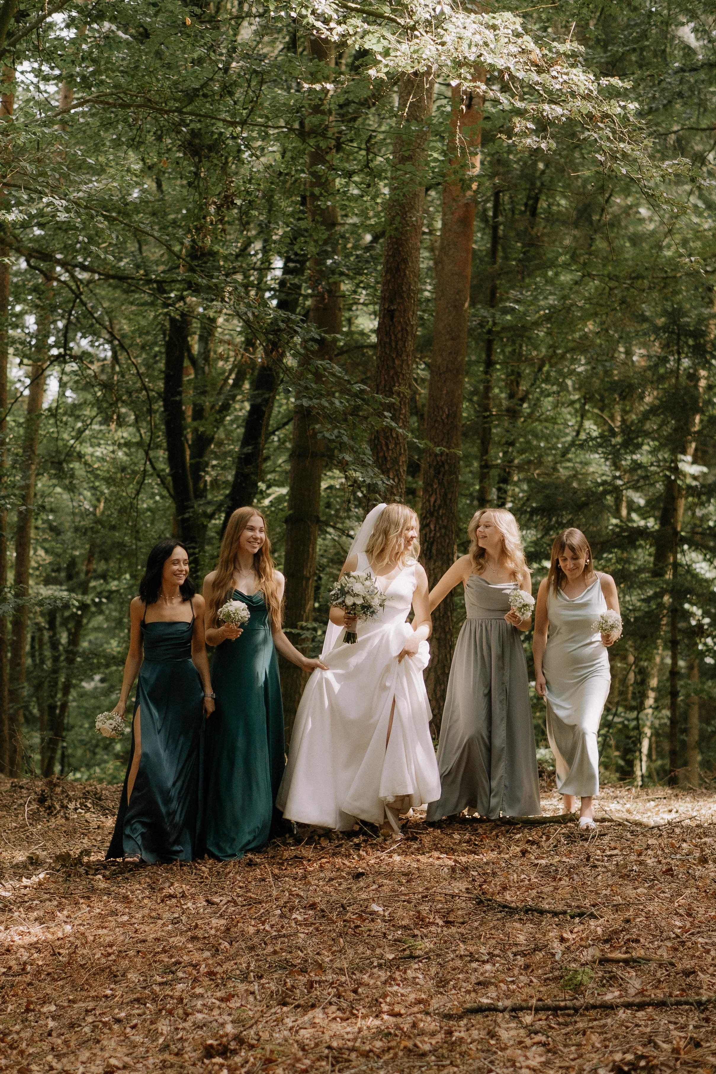 Eine Gruppe von sechs Frauen, darunter eine Braut in einem weißen Brautkleid mit Schleier, die in einem Wald spazieren gehen, umgeben von Bäumen, Blättern und Natur.
