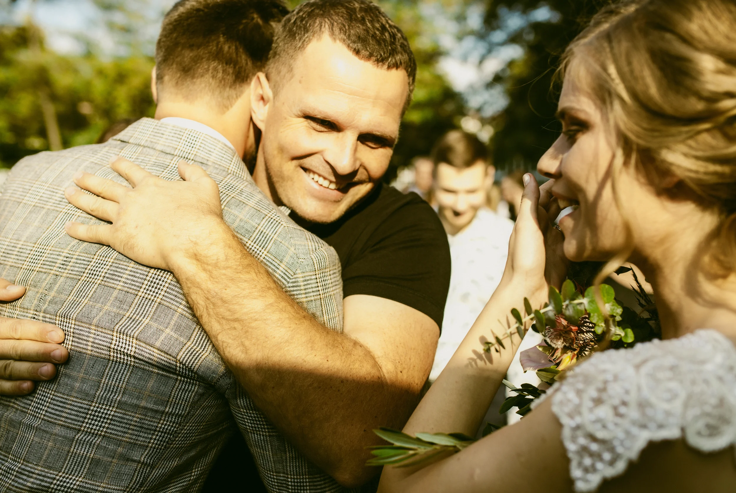 Menschen umarmen sich bei einer Feier im Freien, eine Frau hält einen Blumenstrauß.