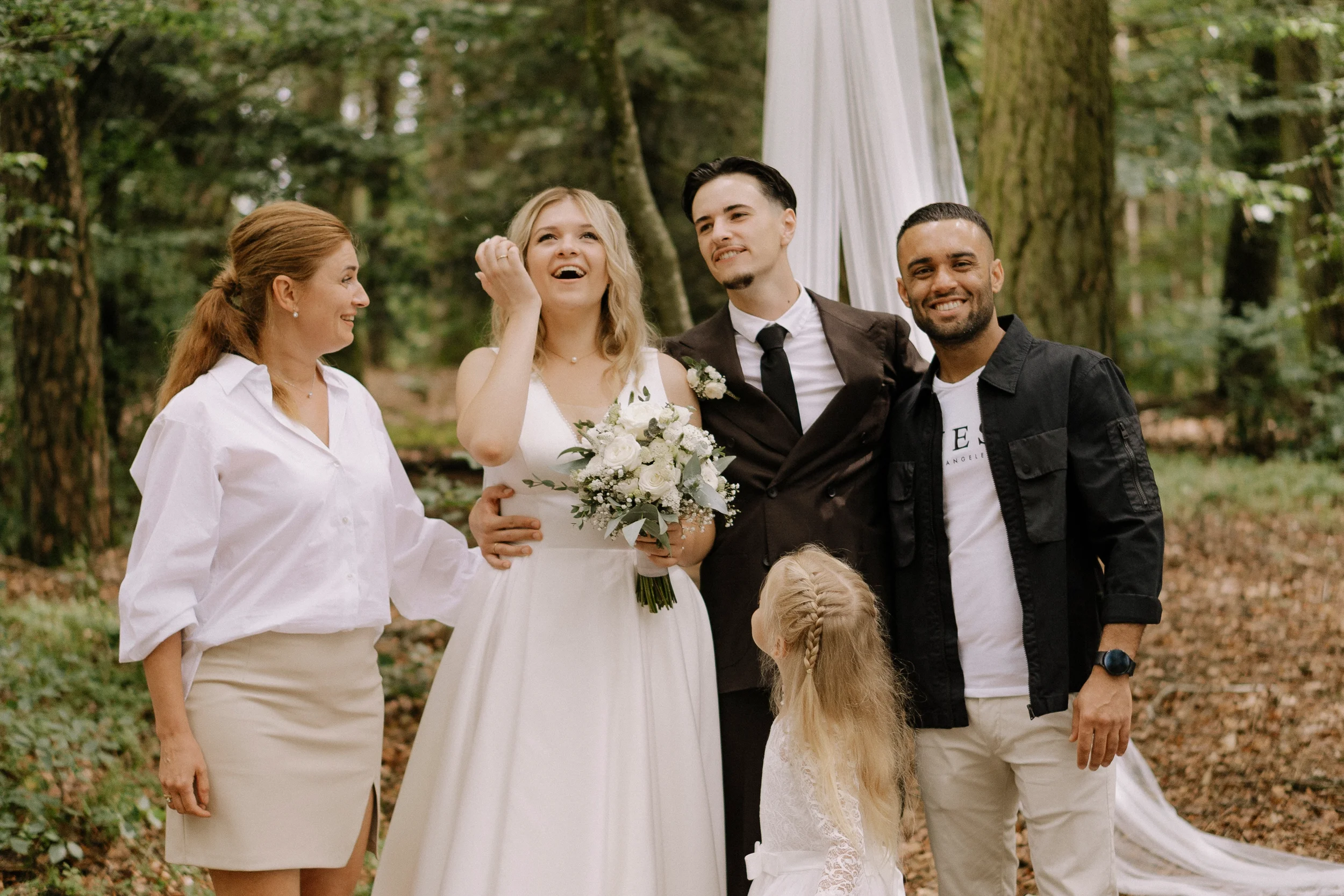 Eine Gruppe von Menschen bei einer Hochzeit im Wald, bestehend aus einem Brautpaar, zwei Frauen und einem Mann, die lachen und sich freuen.