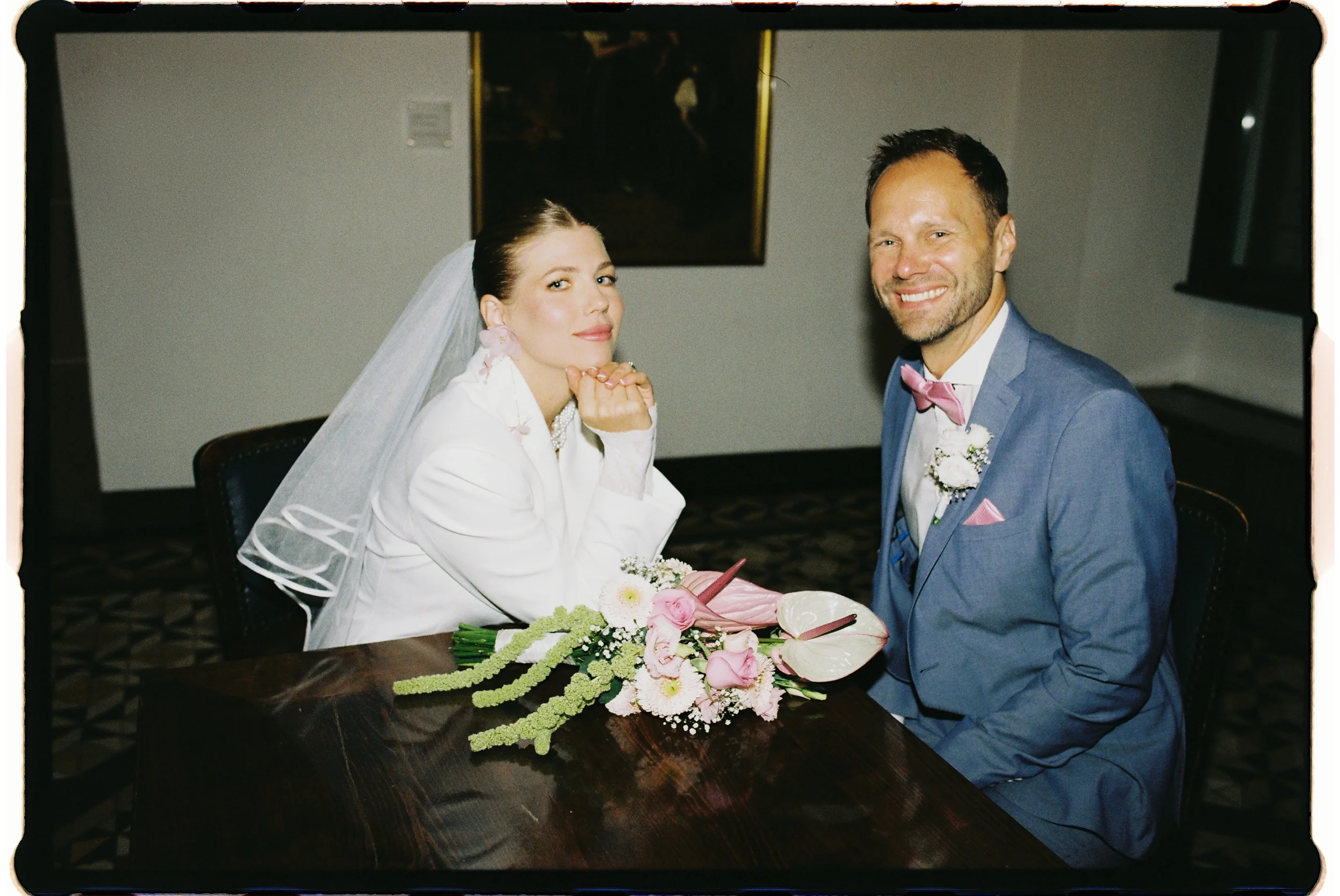 Ein lachendes Brautpaar bei ihrer Hochzeit, die Braut trägt ein weißes Kleid und Veil, der Bräutigam einen blauen Anzug mit rosa Fliege, sie sitzen an einem Tisch mit einem Blumenstrauß.