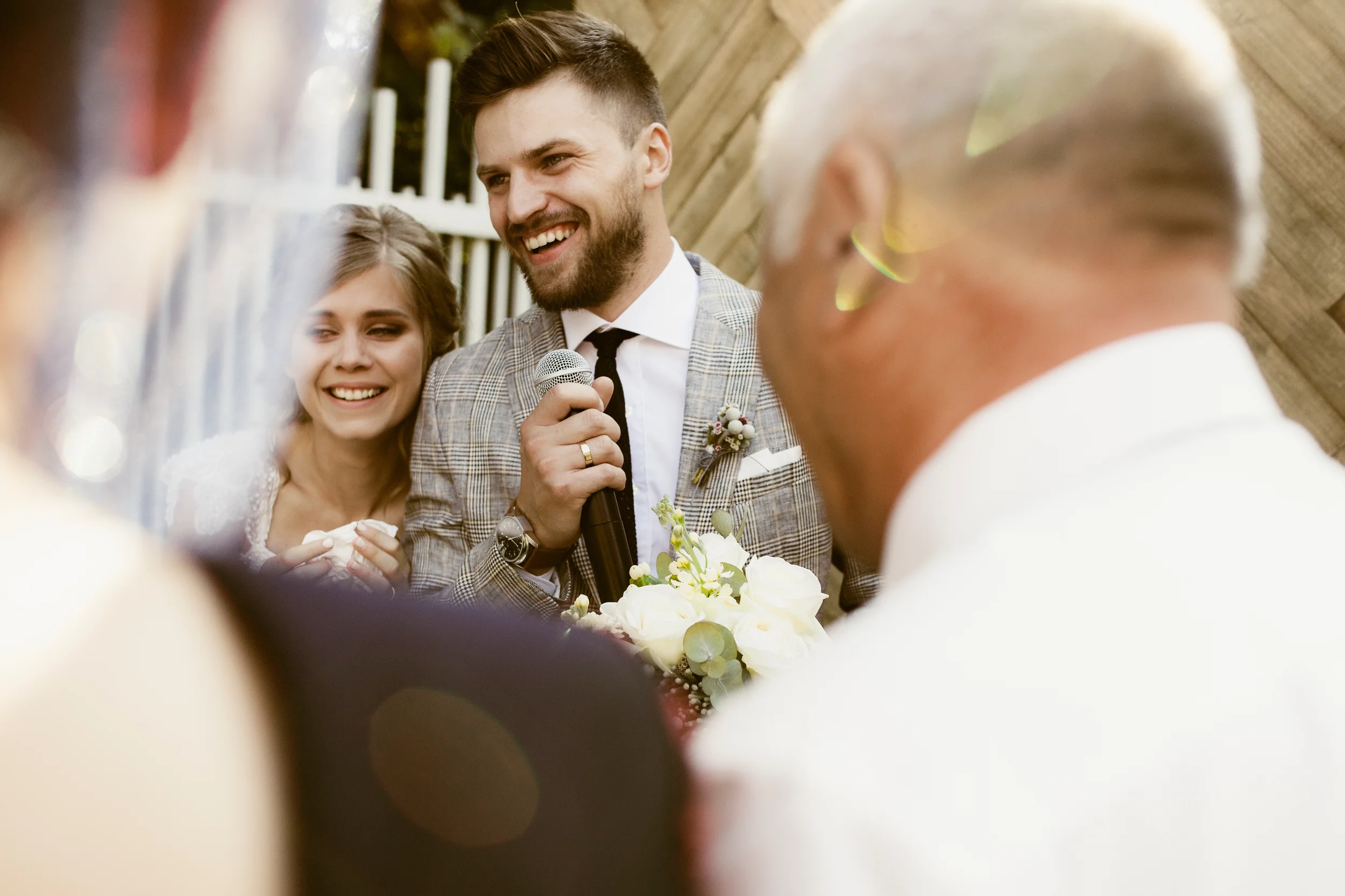 Ein Mann mit Anzug hält eine Mikrofon und lächelt bei einer Hochzeit, umgeben von Braut und anderen Gästen.