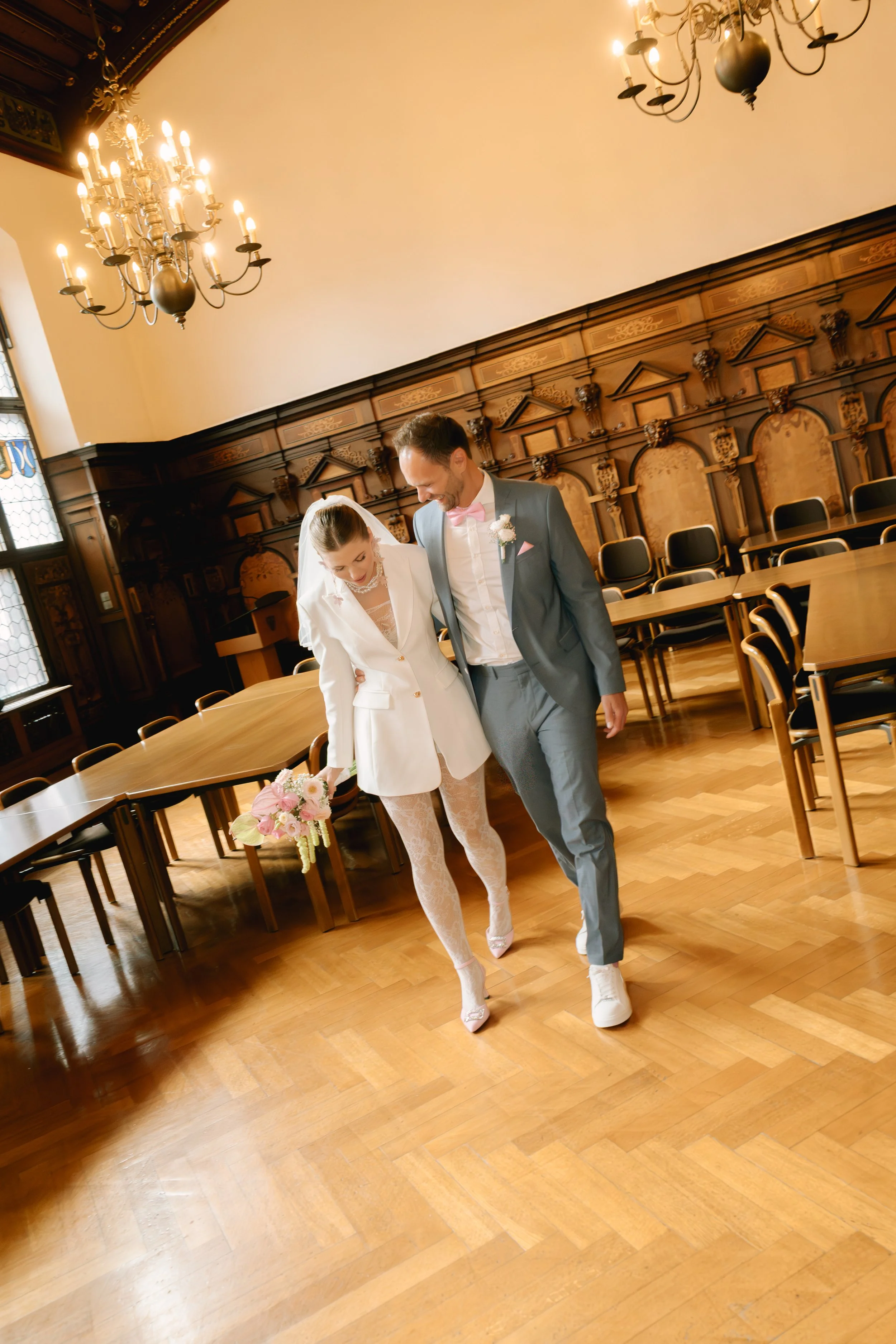 Braut und Bräutigam gehen lachend durch einen eleganten, historisch gestalteten Raum mit Holzvertäfelung und großen Kronleuchtern, teilen Freude und Glück an ihrem Hochzeitstag.