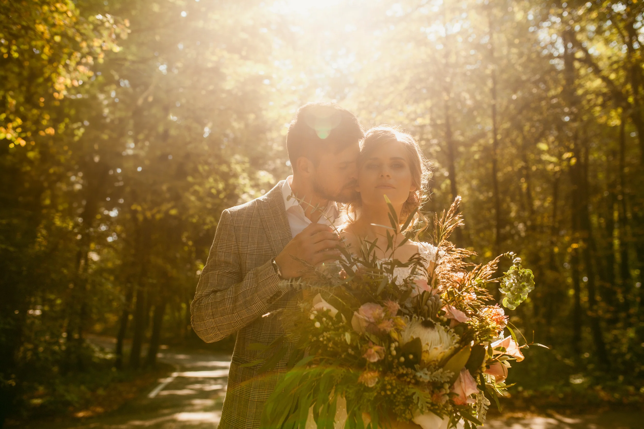 Ein Paar steht im Sonnenlicht in einem Wald, und die Frau hält einen großen Blumenstrauß in den Händen.