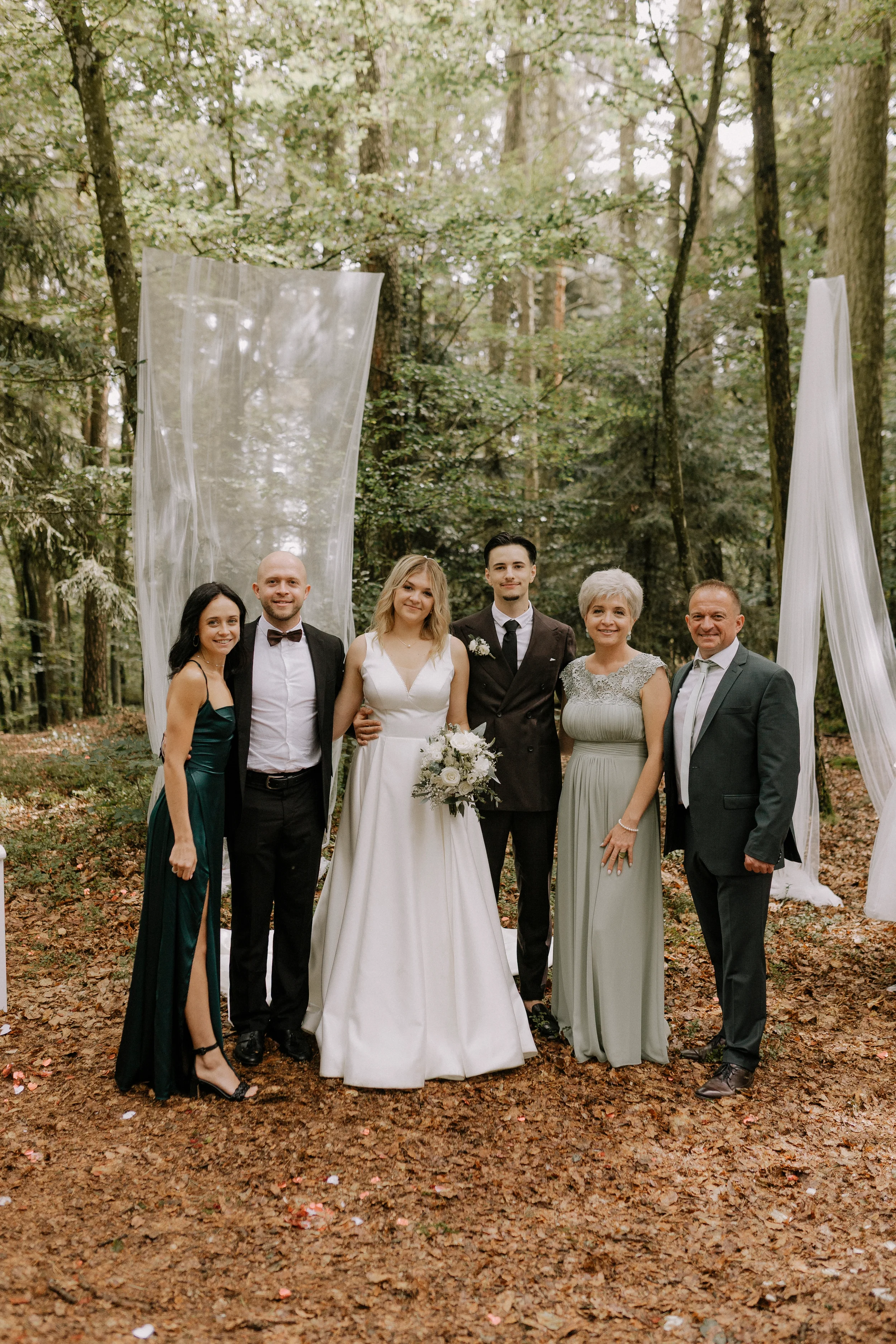 Eine Gruppe von sieben Menschen, bestehend aus Brautpaar und Familienmitgliedern, steht bei einer Hochzeit im Wald.