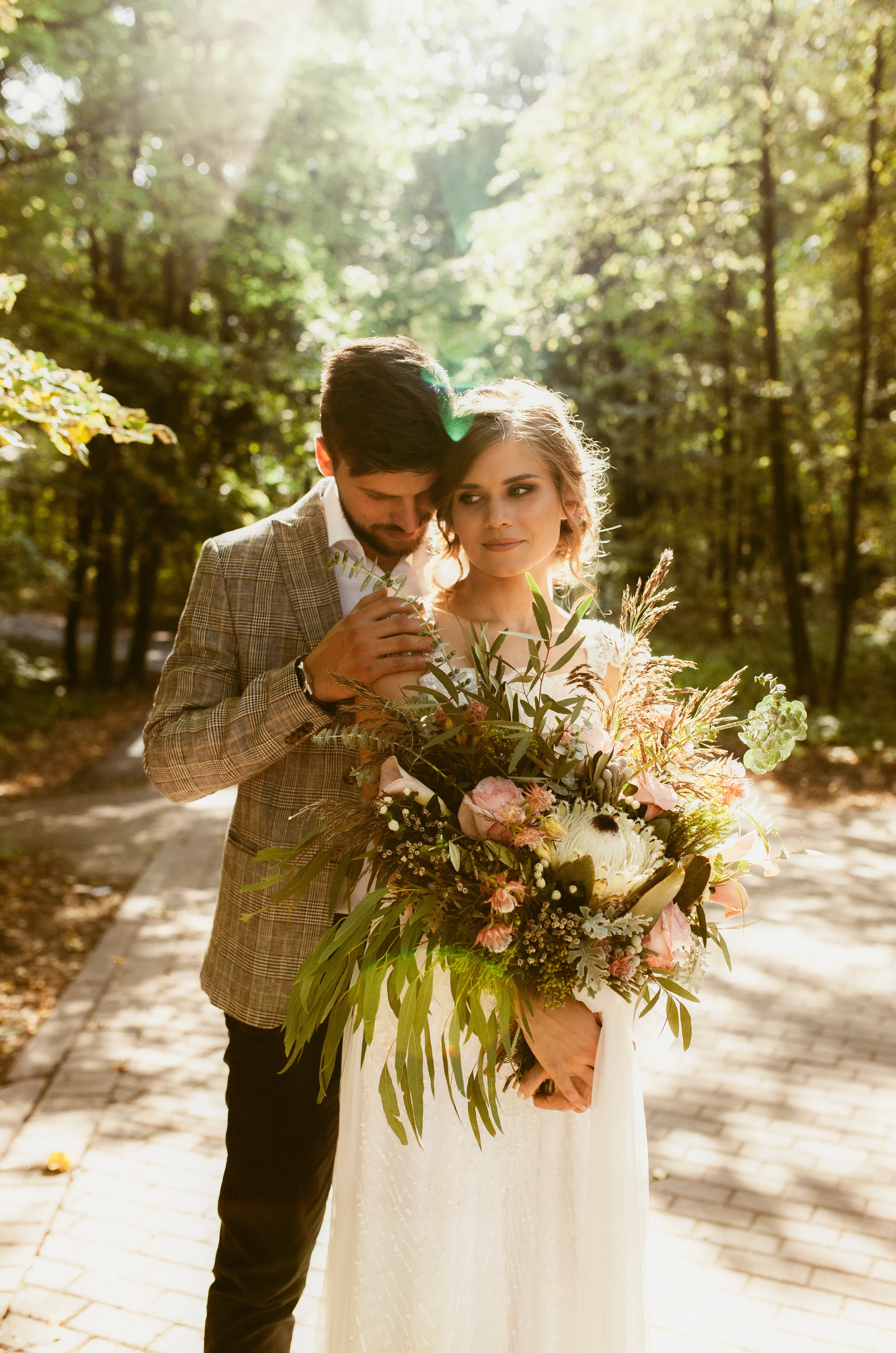 Ein Brautpaar steht im Wald, die Braut hält einen großen Blumenstrauß und trägt ein weißes Hochzeitskleid, während der Bräutigam ein kariertes Jackett trägt.