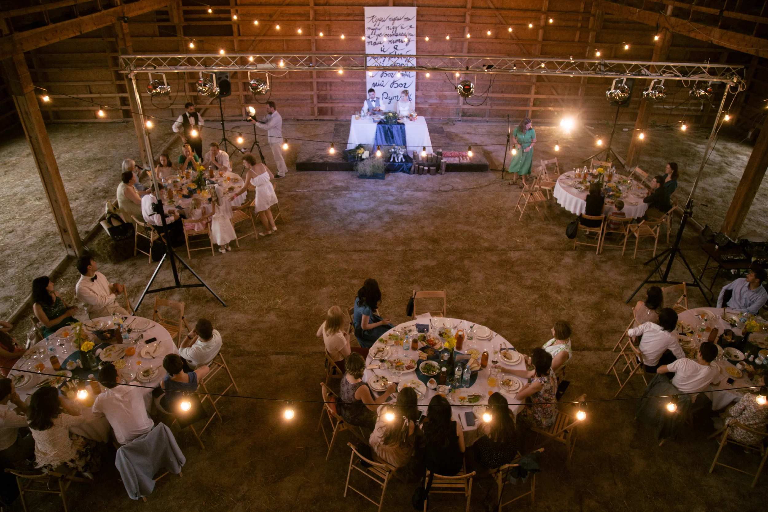 Ein festlicher Raum mit runden Tischen, die mit Blumen und Essen gedeckt sind, und einer Bühne mit DJ und Lichterdekoration in einem rustikalen Scheunengebäude. Es scheint eine Feier oder Hochzeit zu sein.