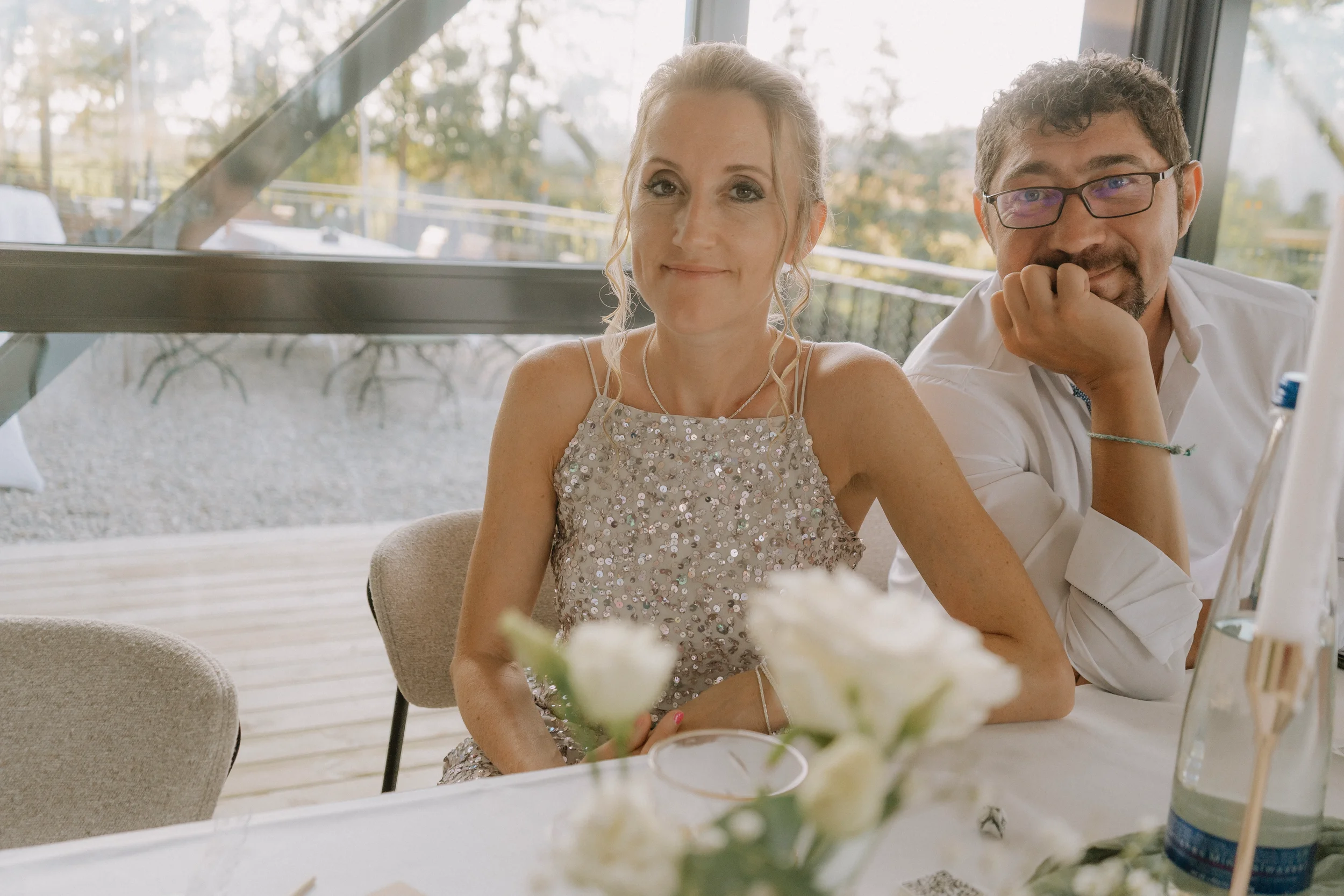 Ein junges Paar bei einem festlichen Anlass, Frau trägt ein silbernes, glitzerndes Kleid, beide sitzen an einem Tisch in einem hellen Raum mit großen Fenstern und Naturblick.