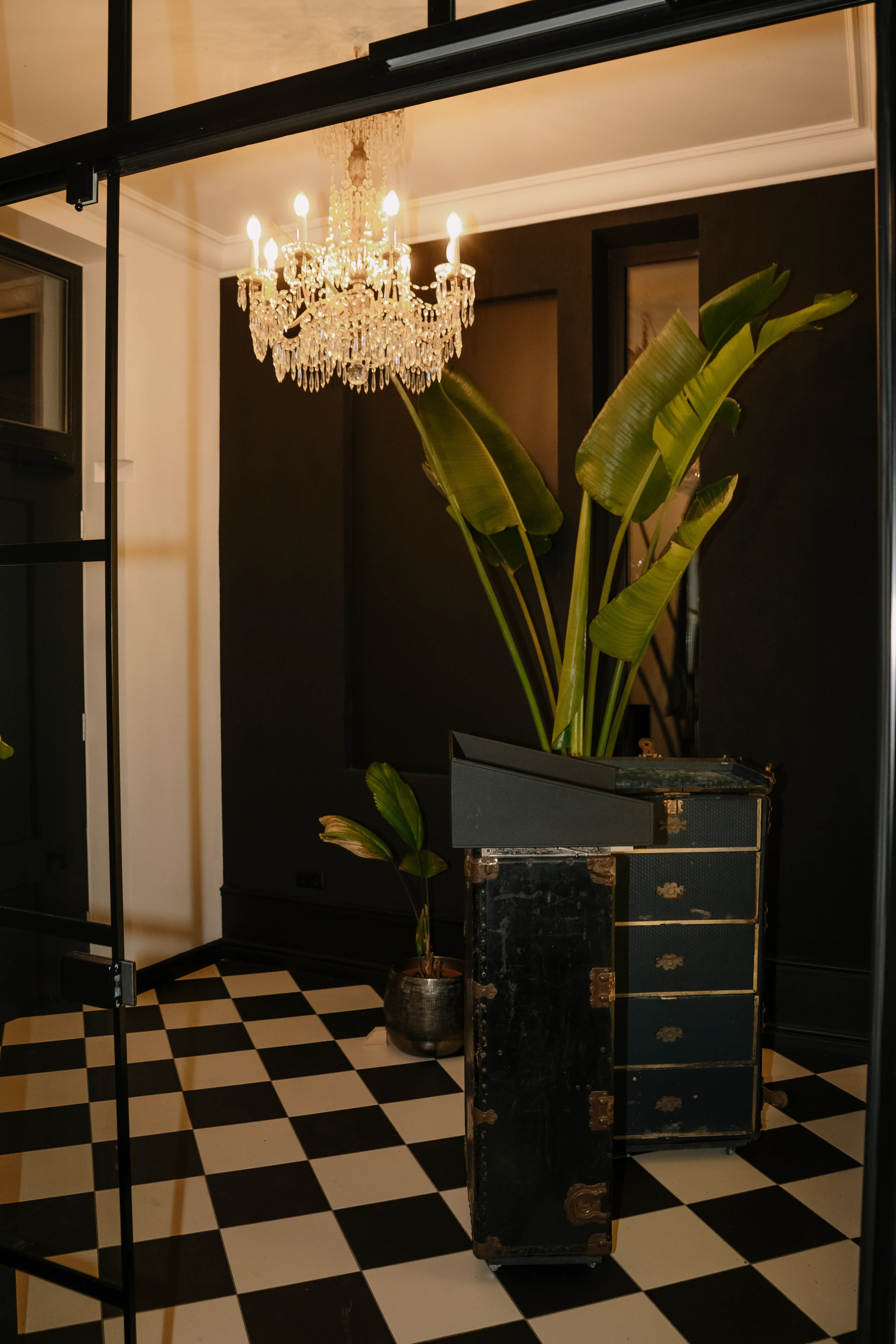 Ein eleganter Raum mit einer Kristallkronleuchte an der Decke, einer großen Pflanze in einem schwarzen Topf und einem schwarzen Koffer mit goldenen Details, das auf einem schwarzen und weißen Fliesenboden steht.