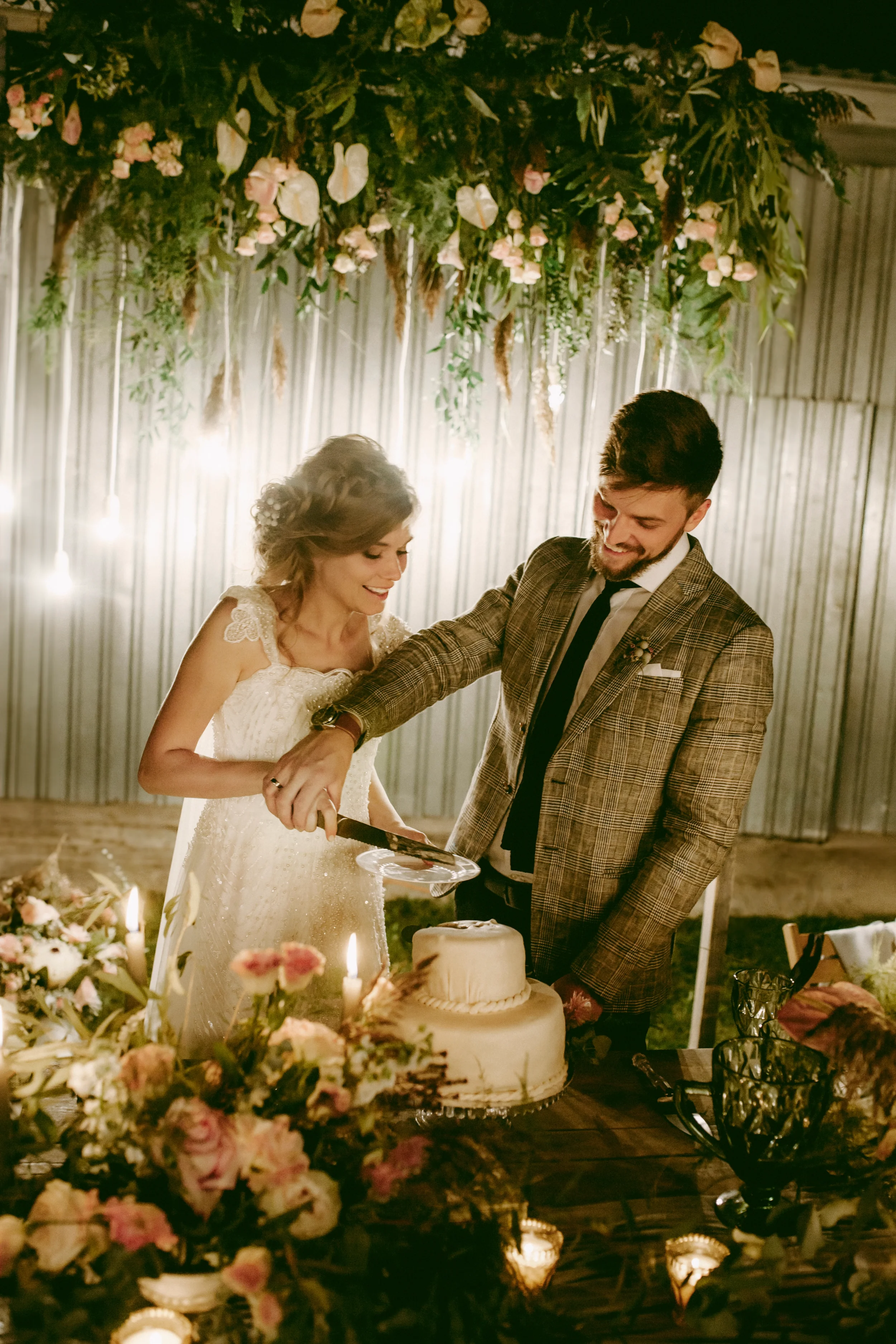 Ein glückliches Paar bei einer Hochzeitstorte, umgeben von Blumendekoration und Kerzen im festlich gedeckten Raum.