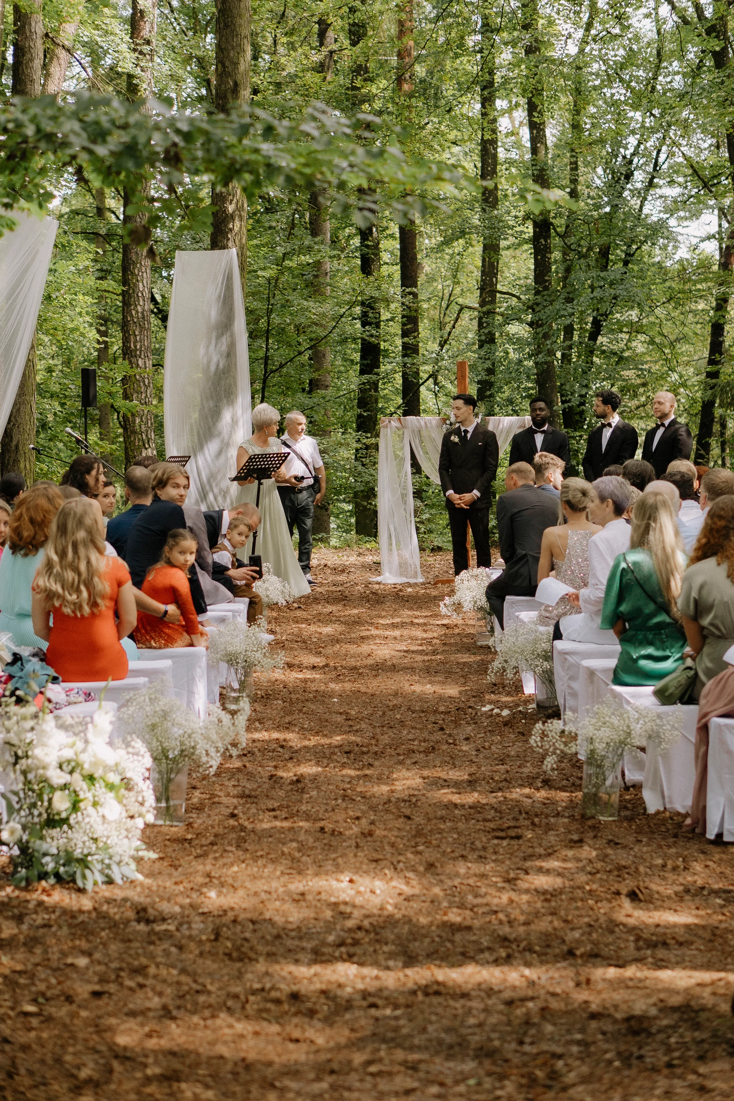 Eine Hochzeitszeremonie im Freien in einem Wald, mit Stühlen, weißen Dekorationen und Gästen, die auf den Bräutigam und die Braut warten.