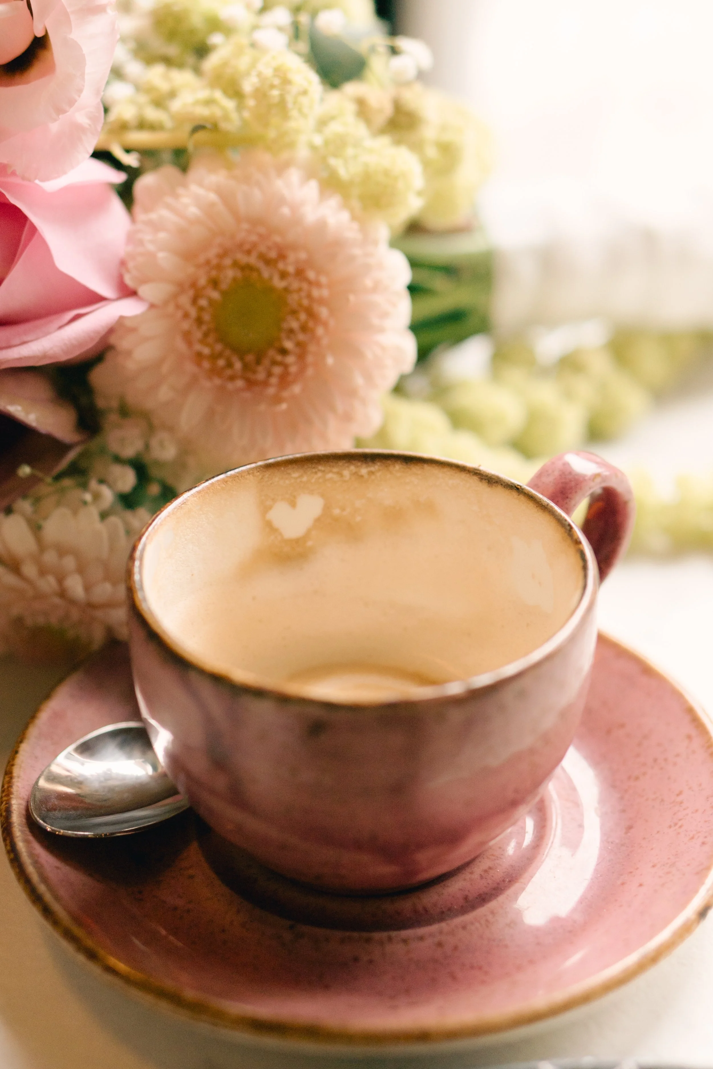 Eine rosa Tasse mit Kaffee oder Cappuccino auf einem passenden Teller, daneben liegt ein Löffel. Im Hintergrund ist eine Blume, vermutlich eine Cosmea, sichtbar.