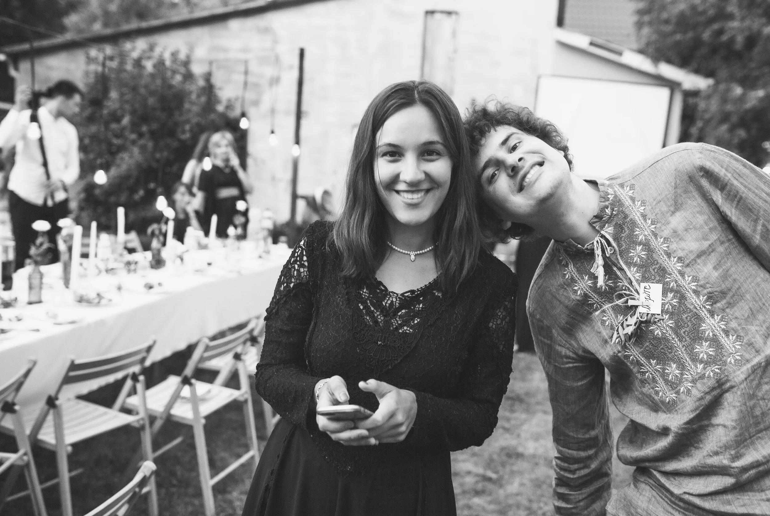 Zwei Menschen, eine Frau mit langem Haar und eine Person mit lockigem Haar, fotografieren Selbstporträt bei einer Feier im Freien, im Hintergrund Tische mit Essen und Dekoration. Es ist eine festliche Atmosphäre.