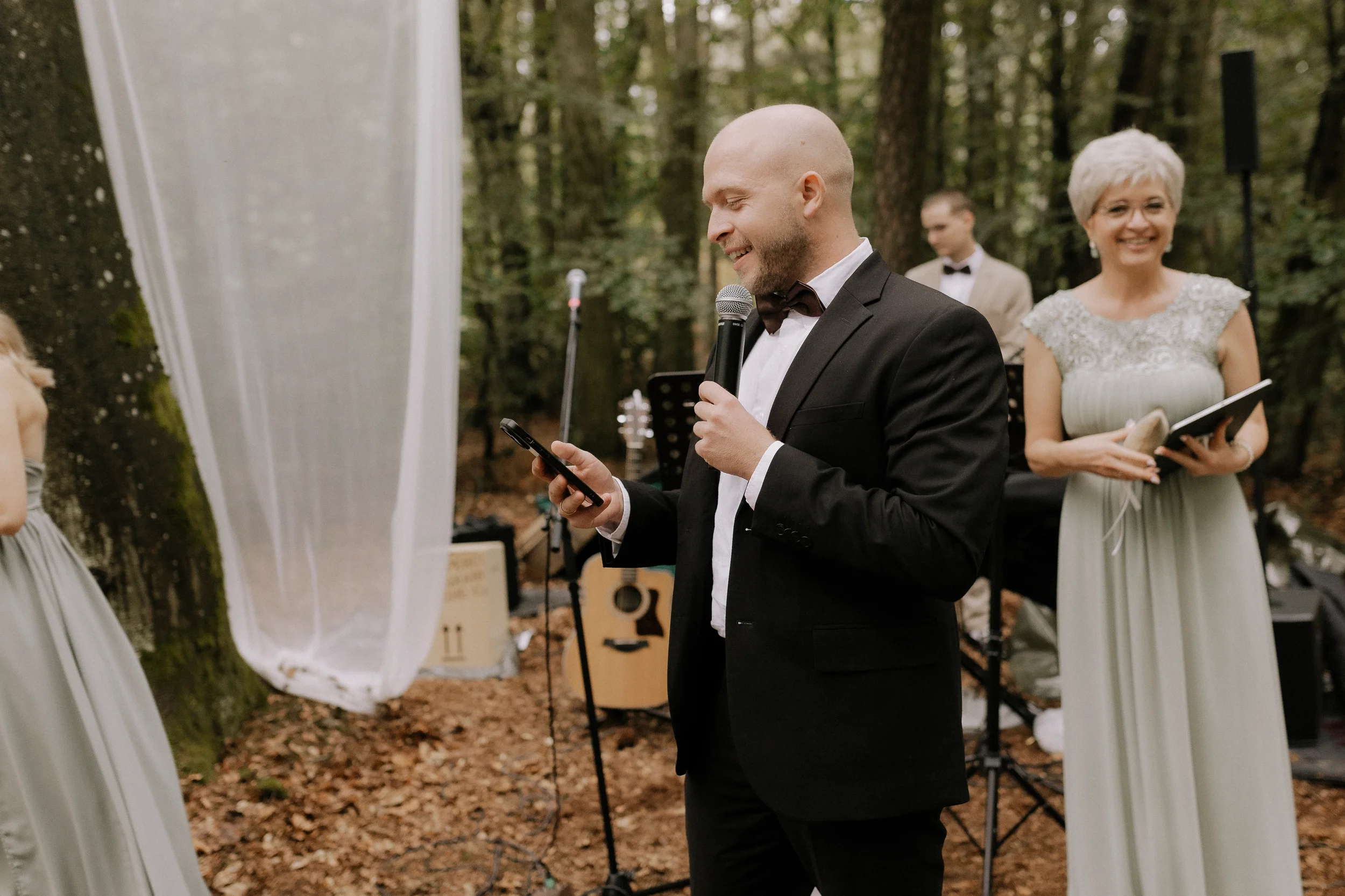 Ein Mann in formeller Kleidung, der bei einer outdoor-Hochzeit eine Rede hält, während im Hintergrund eine Frau in einem weißen Kleid lächelt.