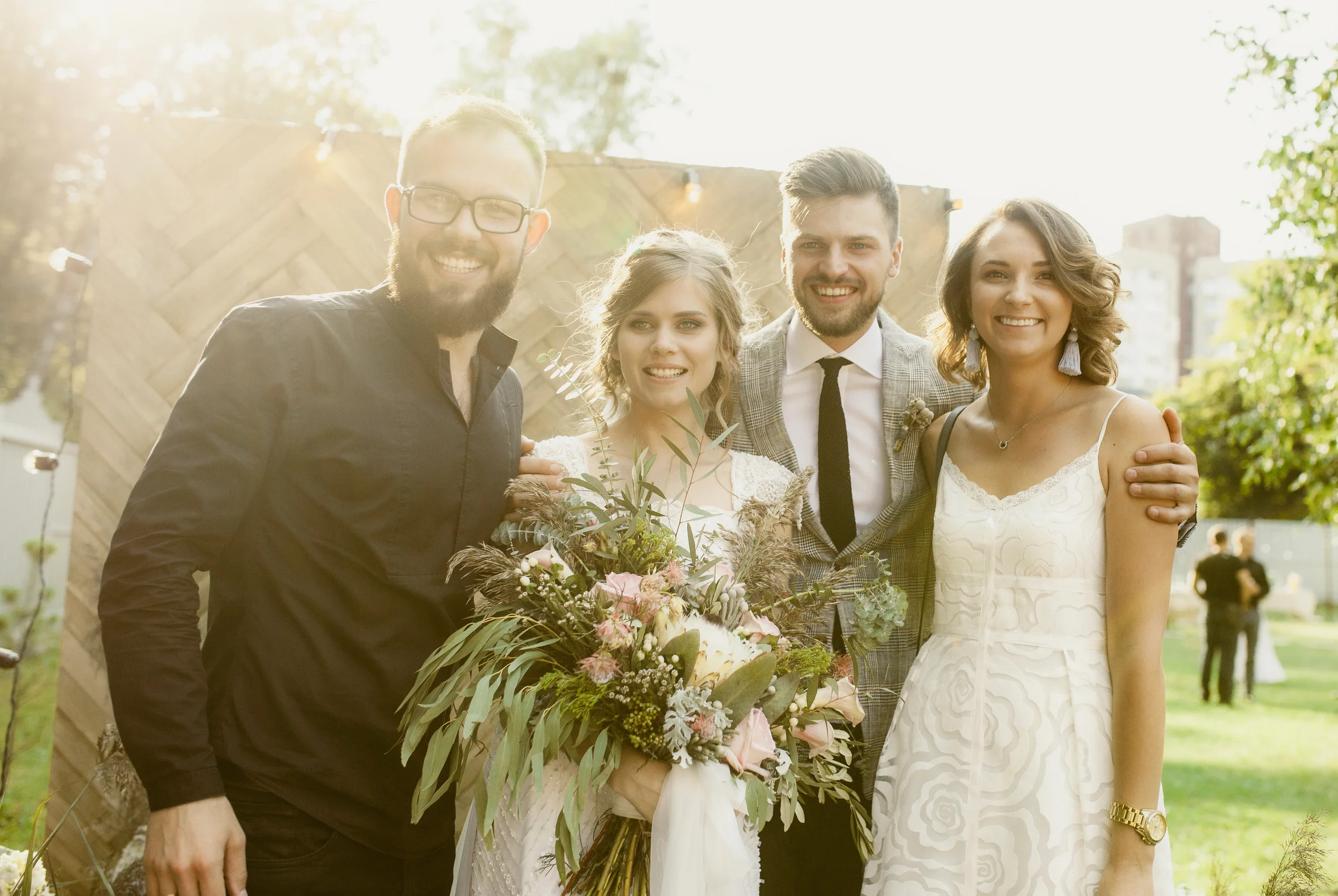 Fünf lachende Menschen bei einer Hochzeit im Freien, umgeben von Bäumen, mit Blumen und dekorativen Lichtern im Hintergrund, sonniges Wetter.