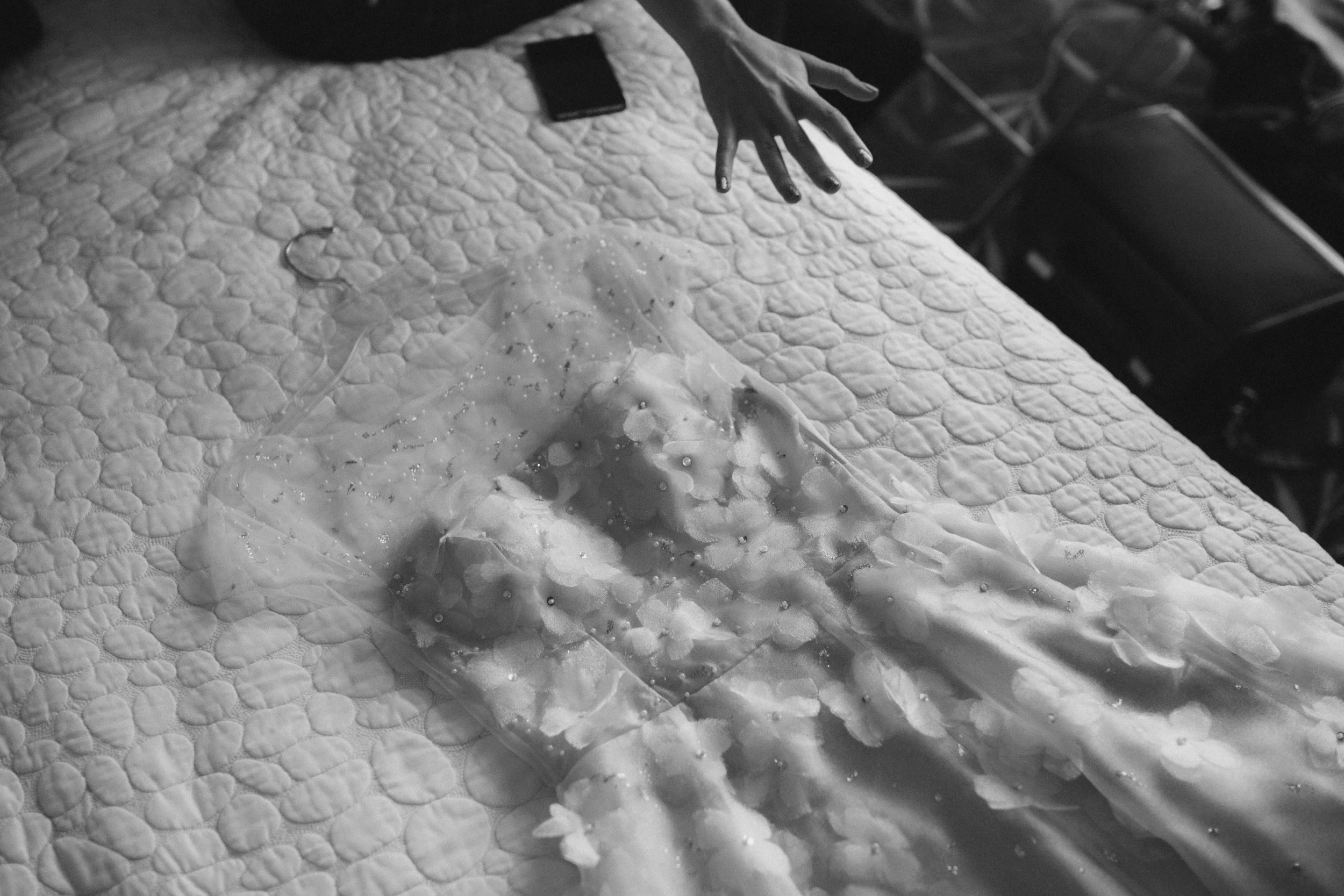 Schwarze Hand, die einen Gegenstand über einer weißen, gezopften Textiloberfläche hält. Das Bild ist in Schwarz-Weiß und zeigt einen Stoff, auf dem eine Plastiktüte mit Perlen oder kleinen Gegenständen liegt.