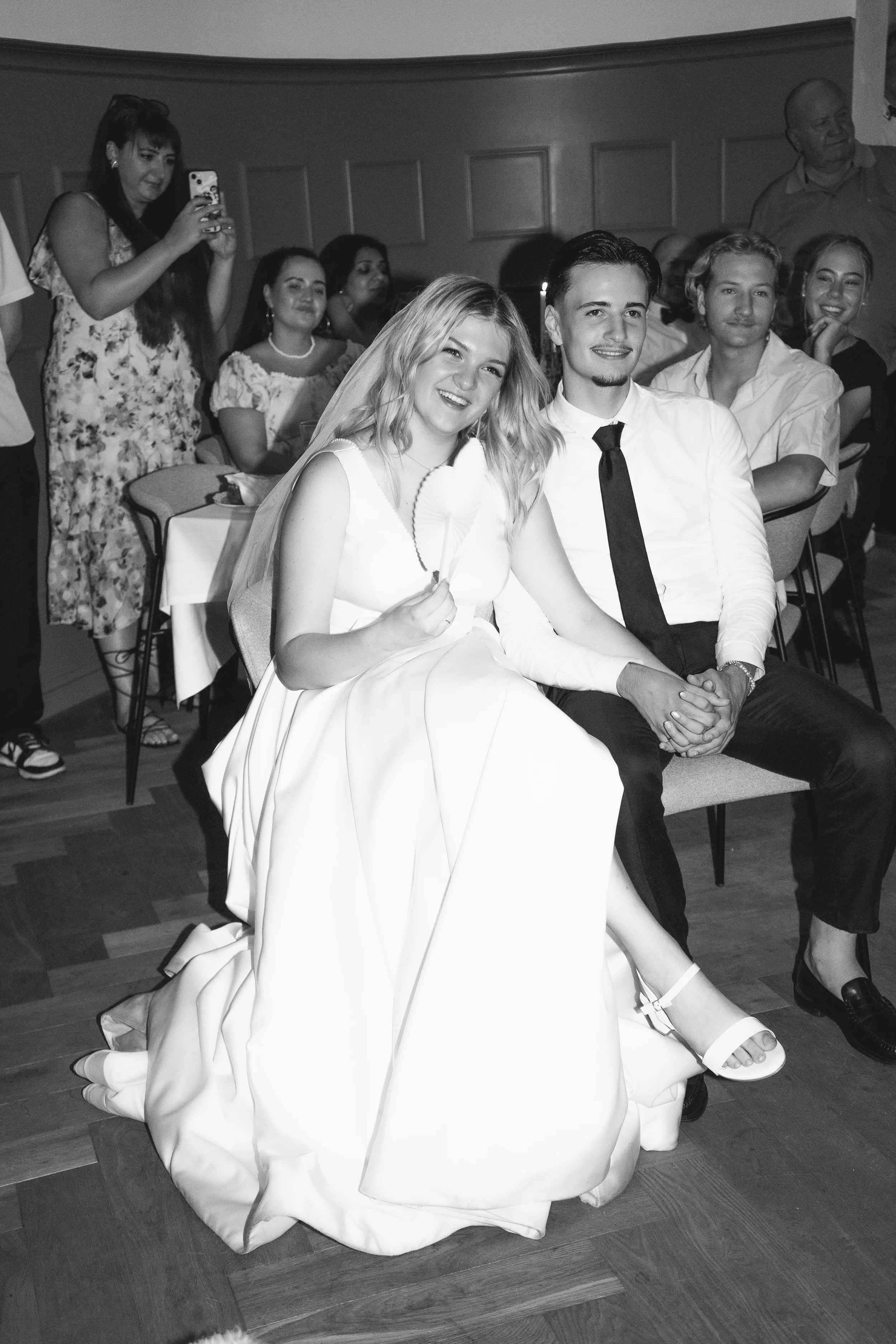 Ein Brautpaar, die Braut in einem weißen Brautkleid und der Bräutigam in einem weißen Hemd mit schwarzen Krawatte, sitzt zusammen, während eine Gruppe von Gästen im Hintergrund zuschaut und Fotos macht.