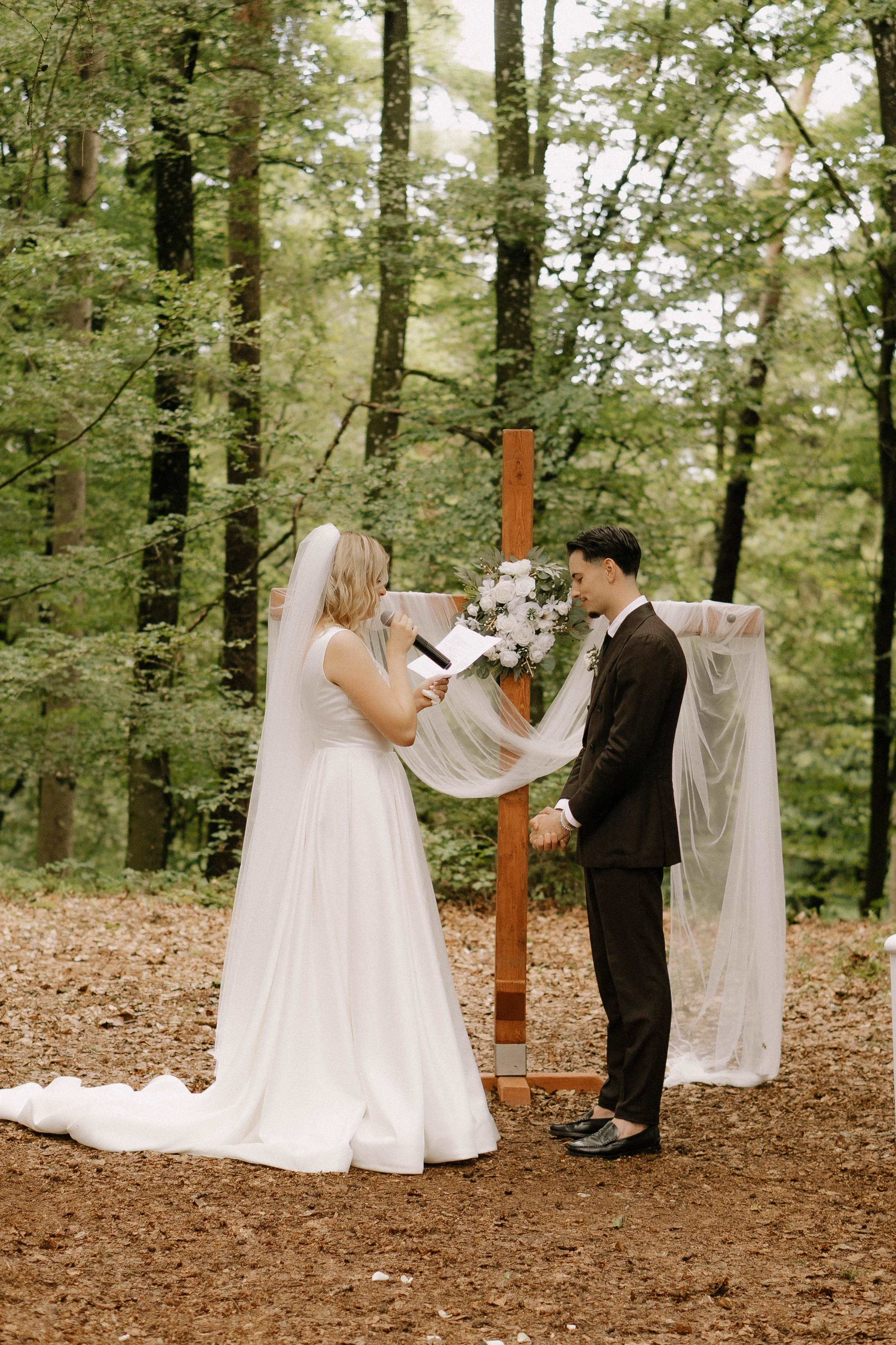 Ein Brautpaar bei einer Hochzeit in einem Wald, die Braut liest gelbe Liebesbriefe vor, der Bräutigam hält ihre Hände, es gibt einen Holzkreuz mit Blumen als Dekoration im Hintergrund.