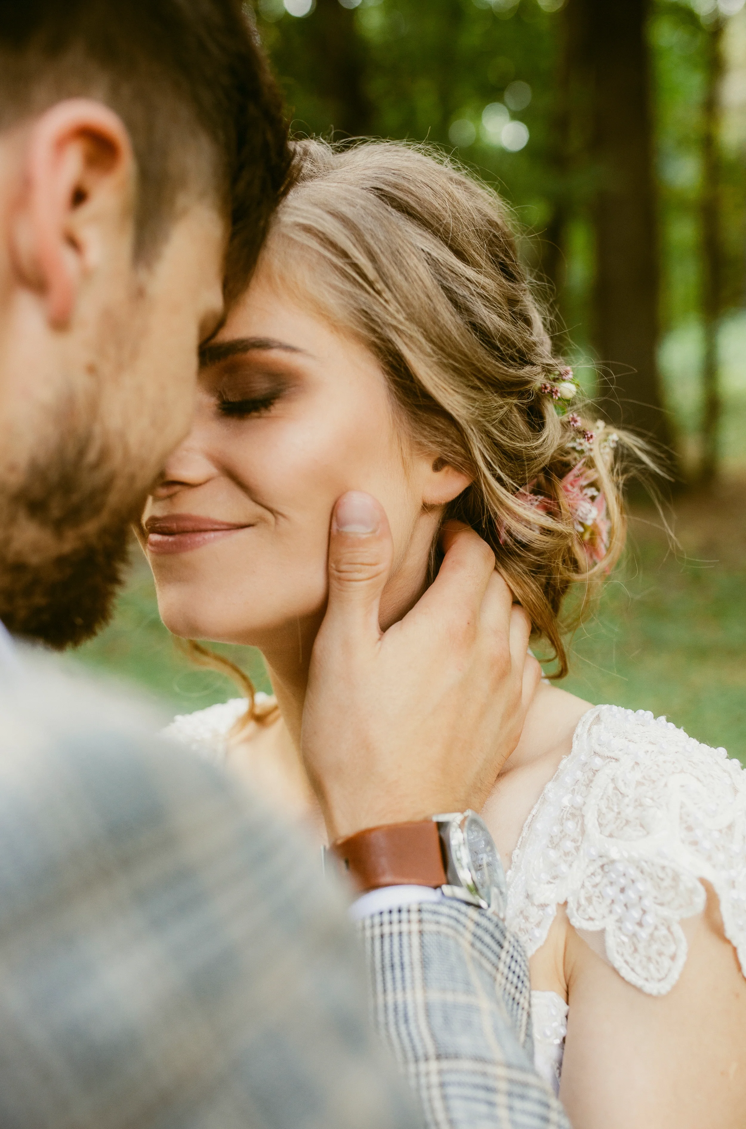 Ein Paar küsst sich zärtlich im Wald, die Frau trägt ein weißes Spitzenkleid, die Männer haben braune Haare, im Hintergrund sind Bäume und grünes Laub.