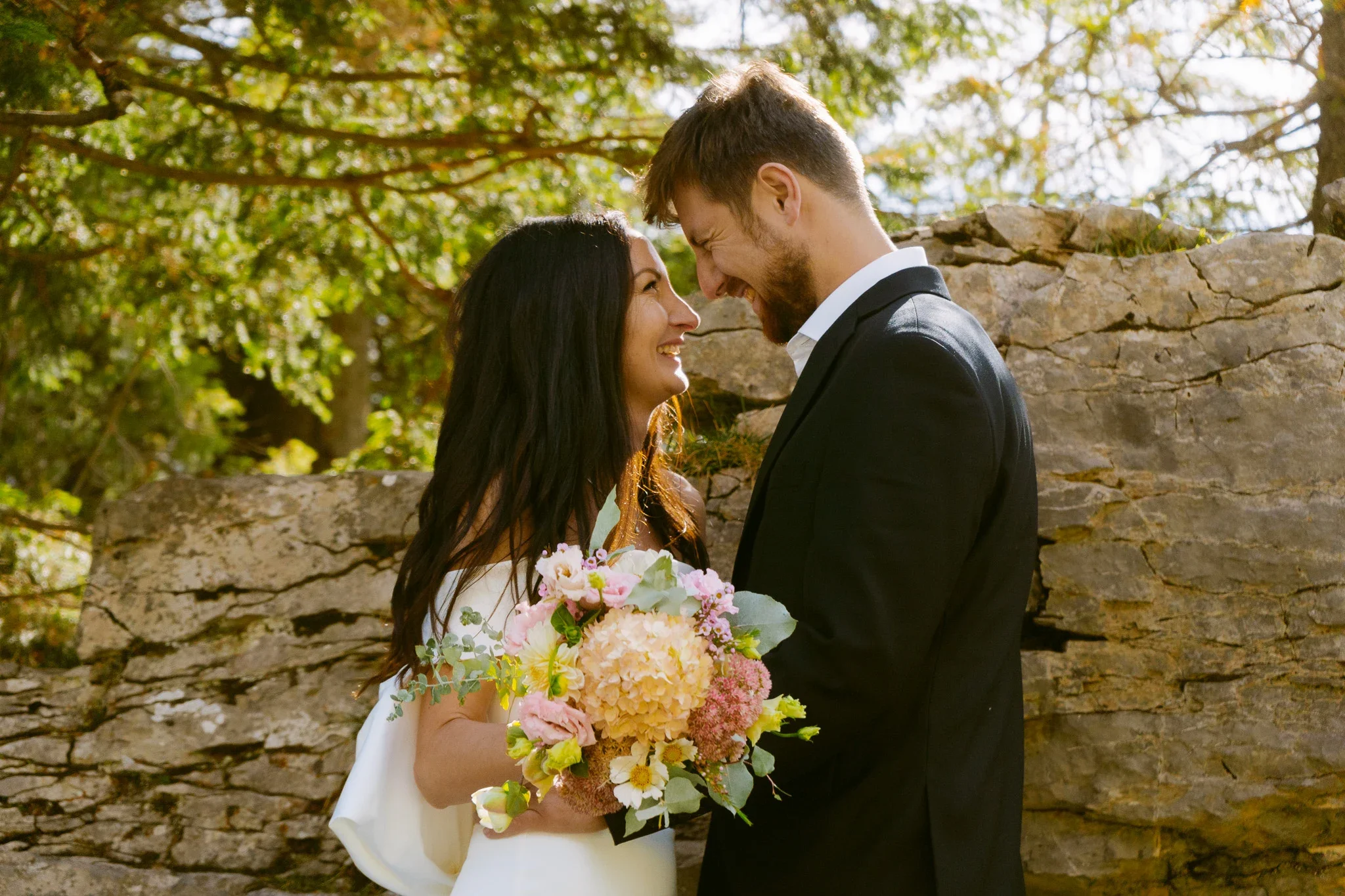 Ein glückliches Paar bei einer Hochzeit, steht sich gegenüber, lächelt und berührt die Stirn. Die Braut hält einen großen Blumenstrauß, während sie vor einem Steinmauer umgeben von grünen Bäumen im Freien stehen.