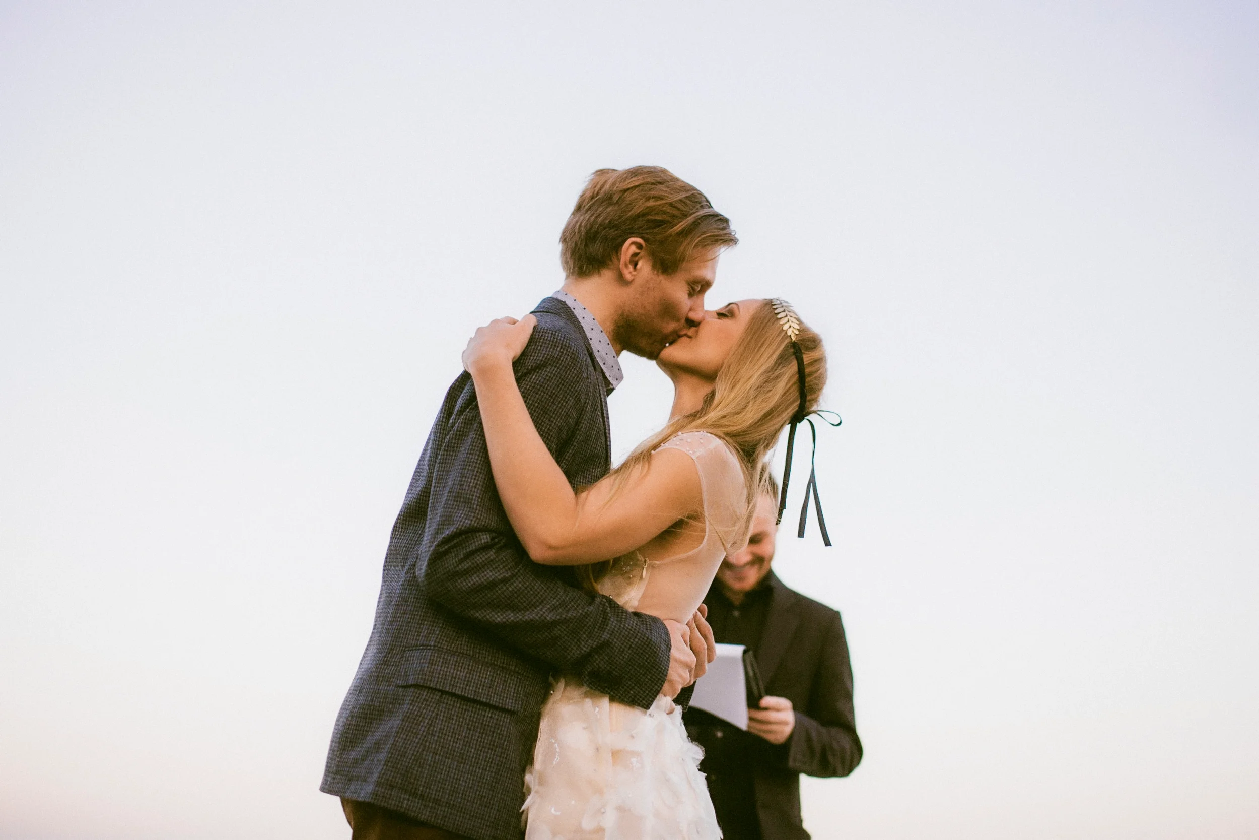 Ein Paar küsst sich bei einer Hochzeit, während ein Mann im Hintergrund liest.