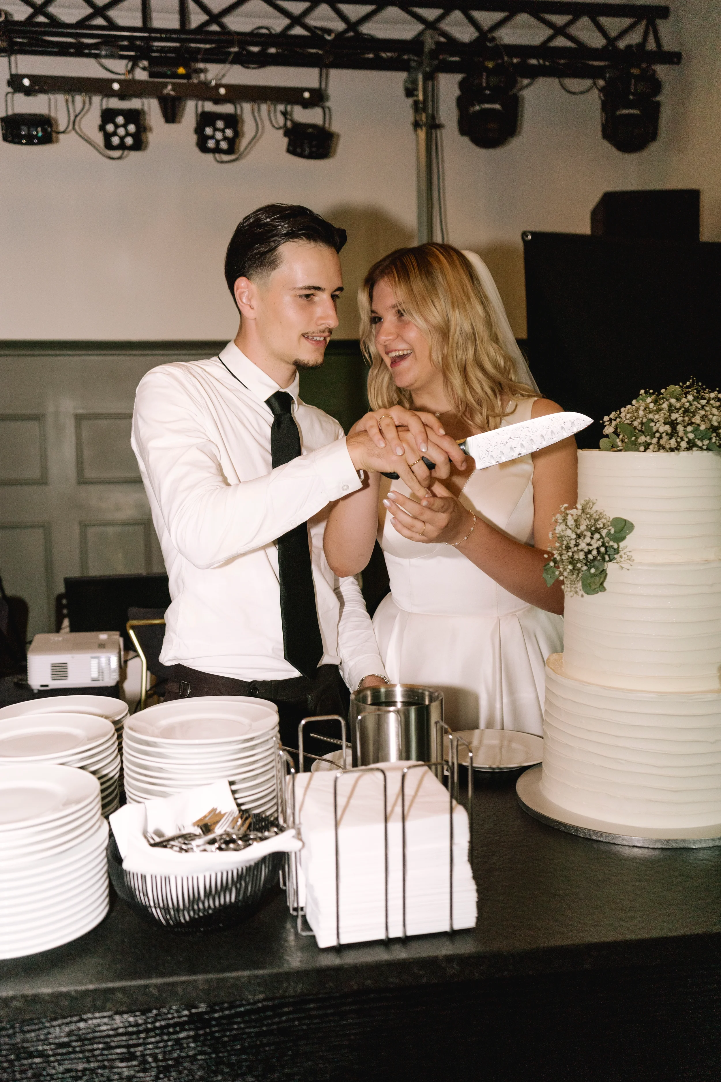 Ein Mann und eine Frau beim Anschneiden einer Hochzeitstorte bei einer Hochzeit im Saal, im Vordergrund sind Geschirr, Besteck und Servietten auf einem Tisch sichtbar.
