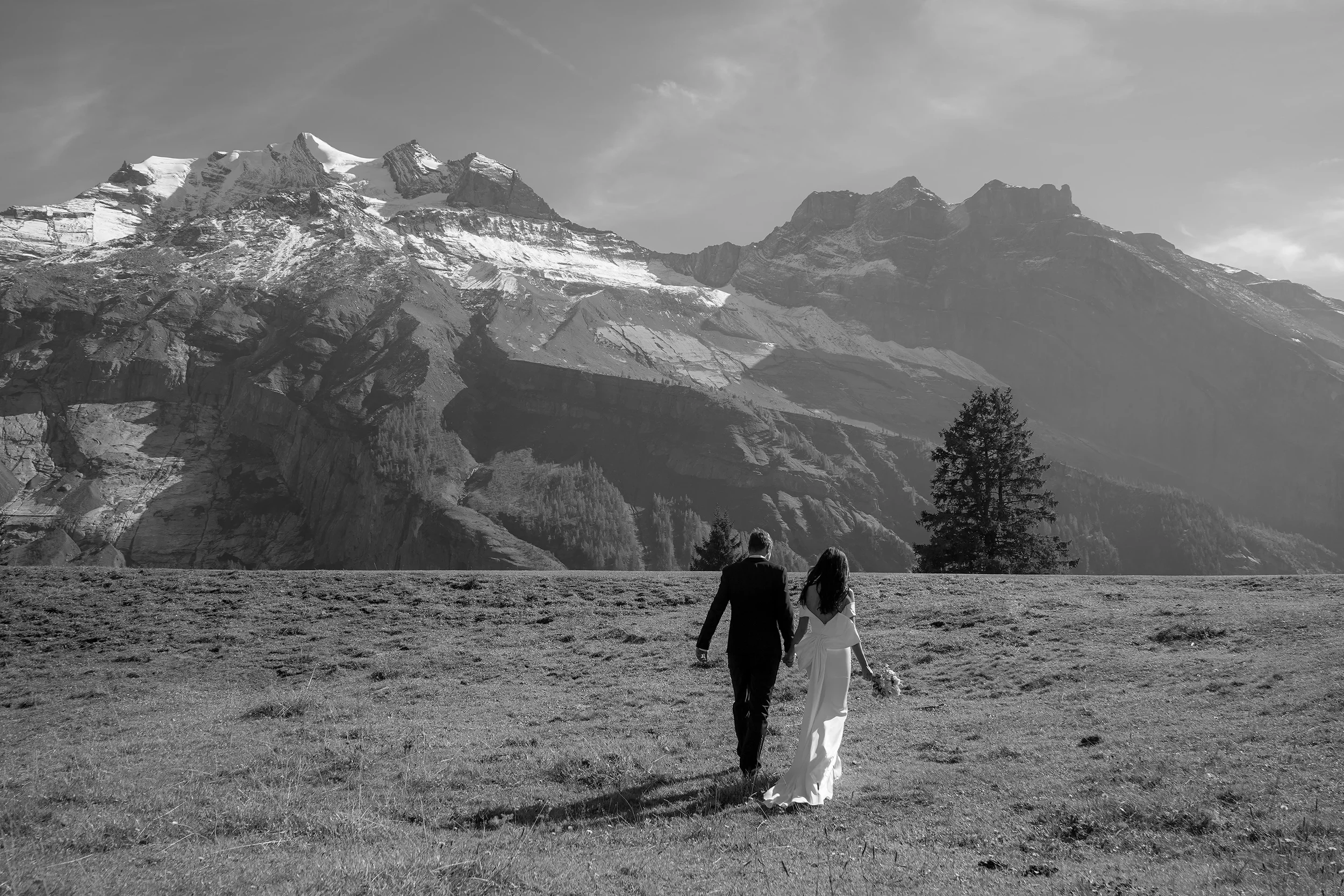 Ein Brautpaar, das Hand in Hand auf einer Wiese vor majestätischen, schneebedeckten Bergen steht, in Schwarz-Weiß.