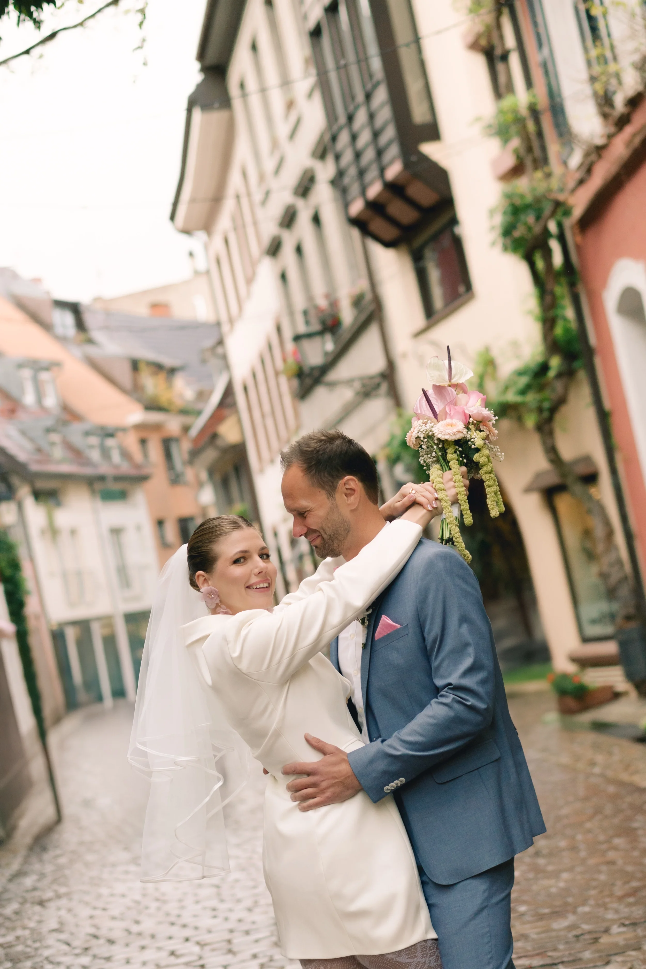 Ein frisch verheiratetes Paar tanzt glücklich auf einer Kopfsteinpflasterstraße in einer Altstadt mit bunten Fassaden und Balkonen mit Blumen. Die Frau trägt ein weißes Kleid, der Mann einen blauen Anzug.