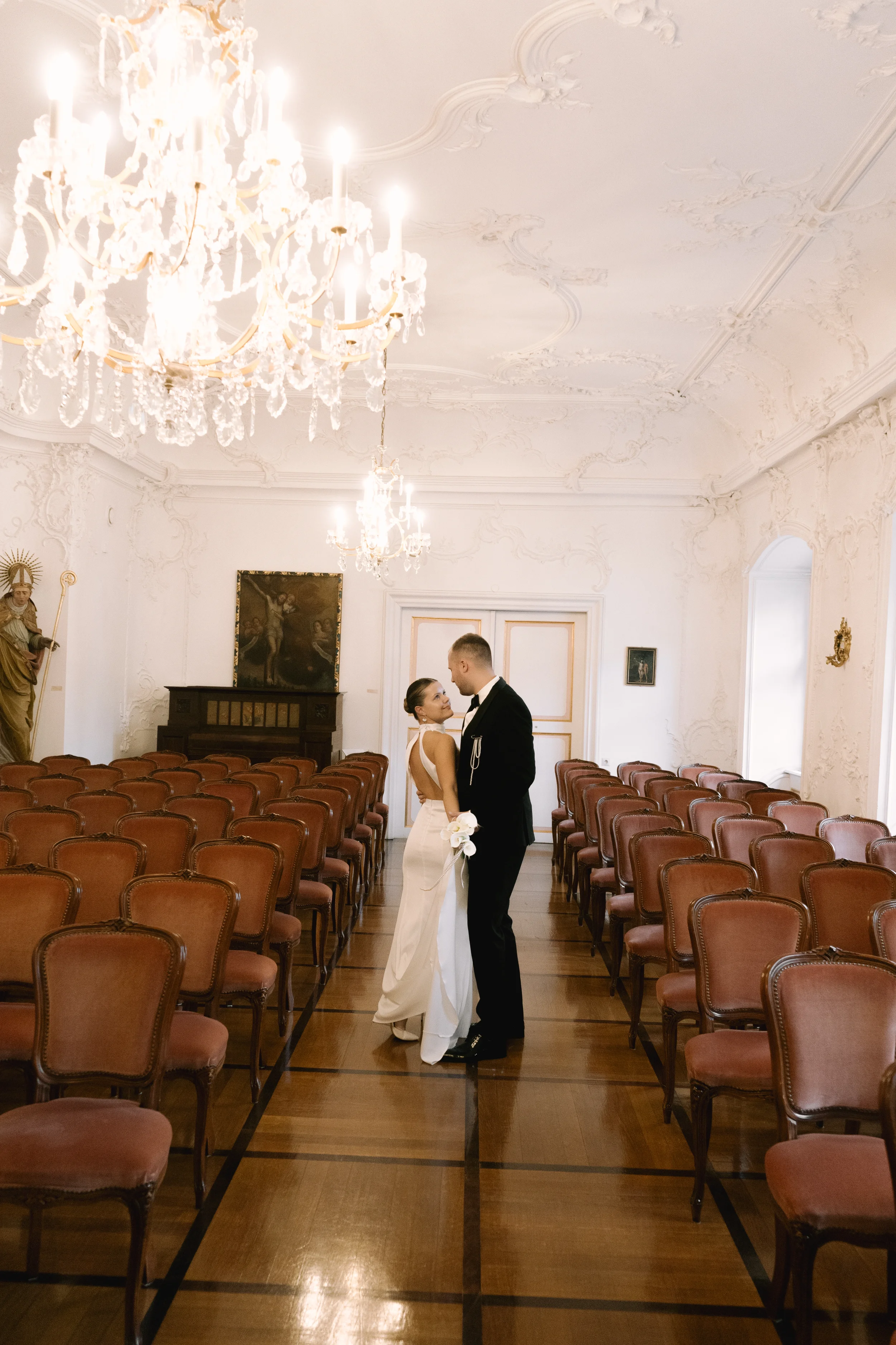 Ein braunes Haar junges Paar in Hochzeitsshirt, das sich im Saal eines elegant eingerichteten Raums mit weißen Wänden und Kronleuchtern gegenübersteht, das auf eine romantische Szene hinweist.