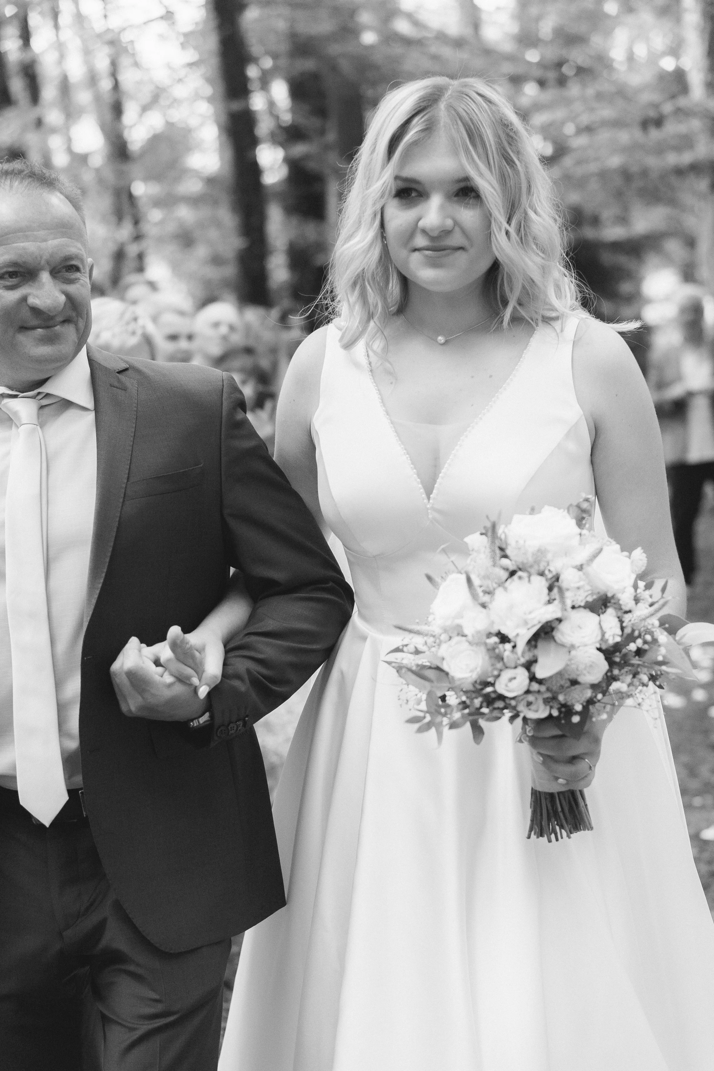 Ein Mann in einem Anzug und eine junge Frau im Brautkleid bei einer Hochzeit im Freien, die gemeinsam die Hand halten.