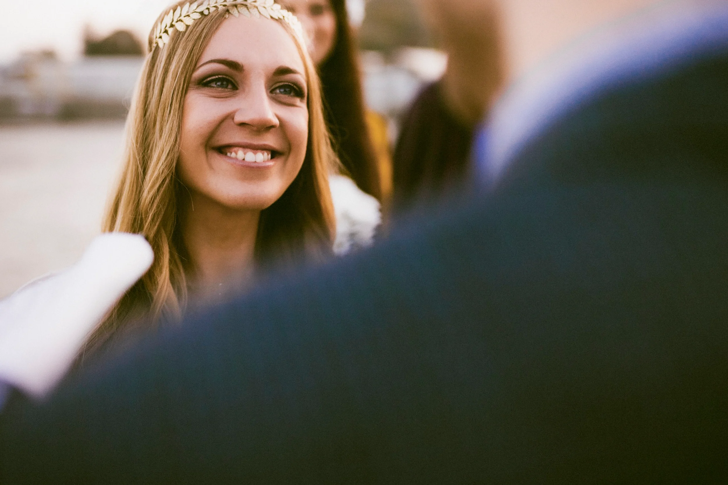 Ein junges Mädchen mit langem, rotem Haar, das ein Blumenkranz trägt, lächelnd und in einem Gespräch mit jemandem, vermutlich bei einer Hochzeit oder festlichen Veranstaltung.