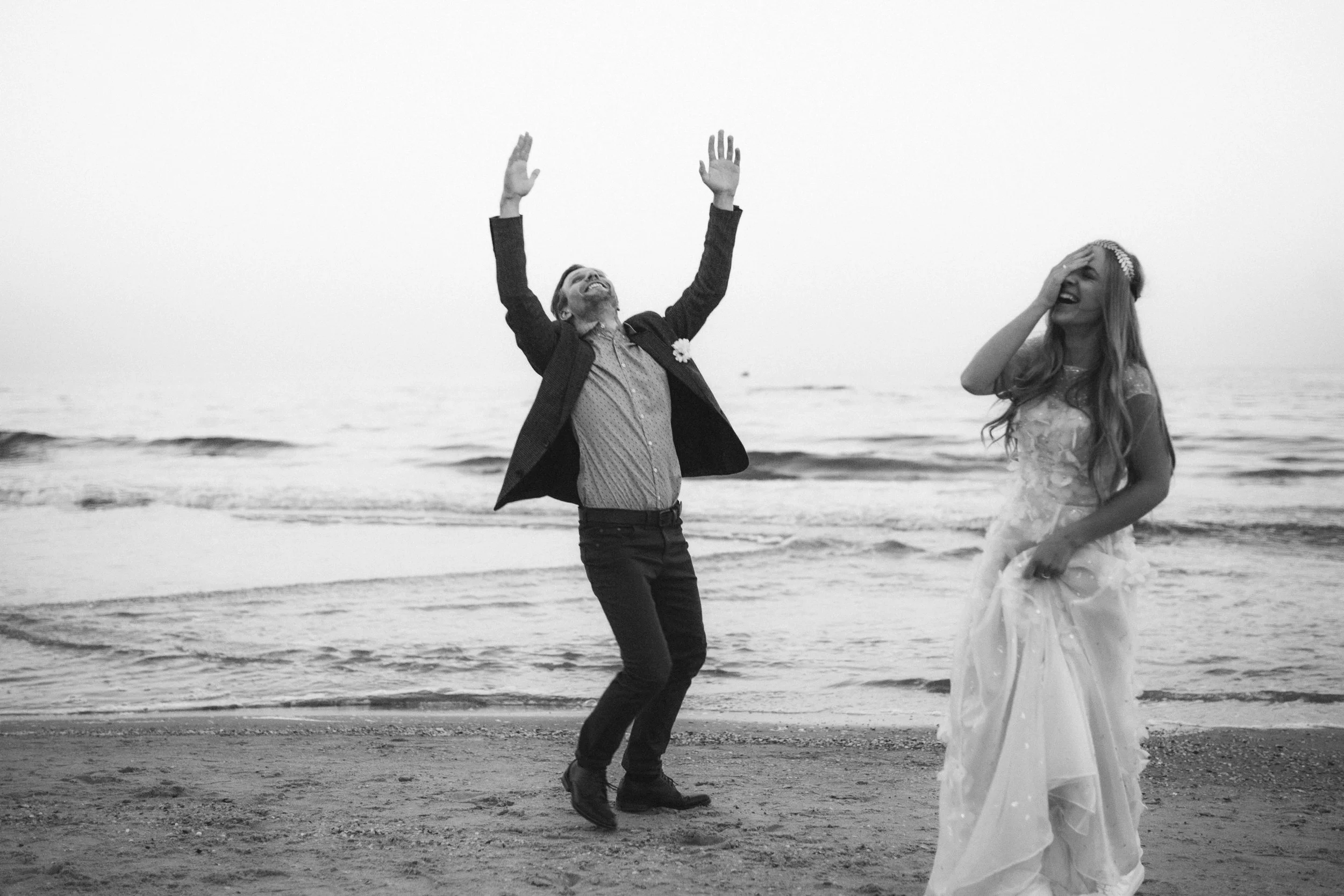 Ein Mann und eine Frau tanzen fröhlich am Strand, der Mann hebt die Arme hoch, während die Frau in einem weißen Brautkleid lächelnd die Hand auf den Kopf legt, im Hintergrund Meer und Himmel.